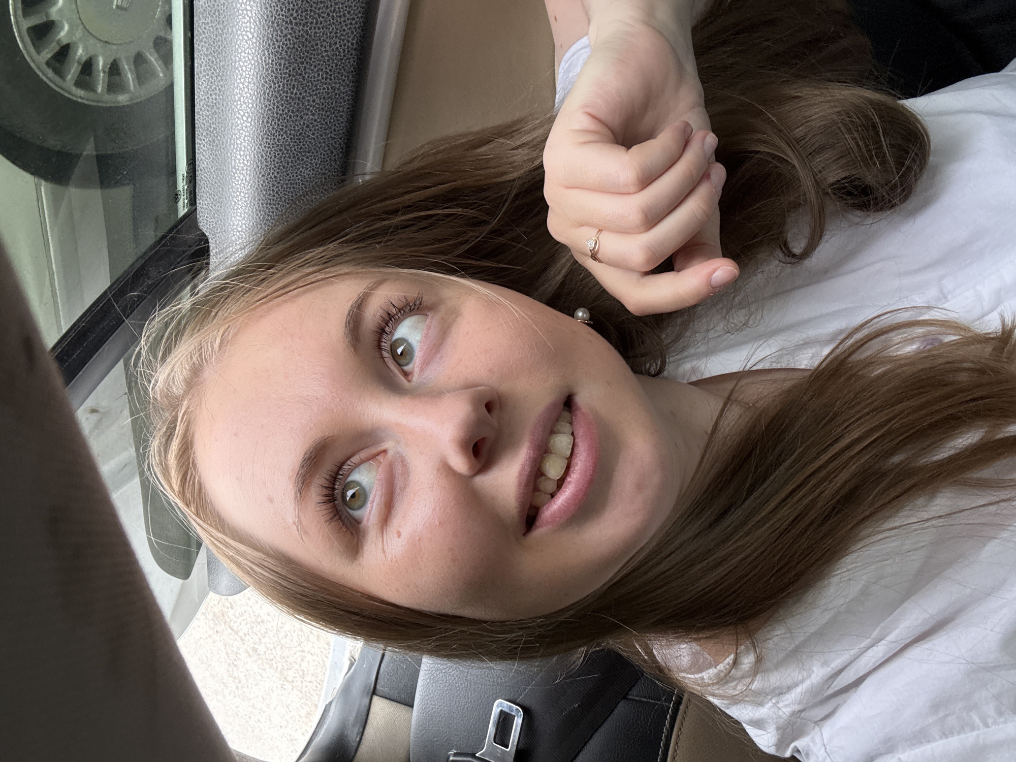 The image shows a young woman from the chest up, looking to her left. She has long, light brown hair and is wearing a white shirt. Her left hand is raised to her face, and she is wearing a ring on her left ring finger. She appears to be in a car, with a seatbelt buckle visible to her left and a car tire visible through the window behind her. The background is out of focus, but appears to be a street scene. The overall lighting suggests it is daytime.