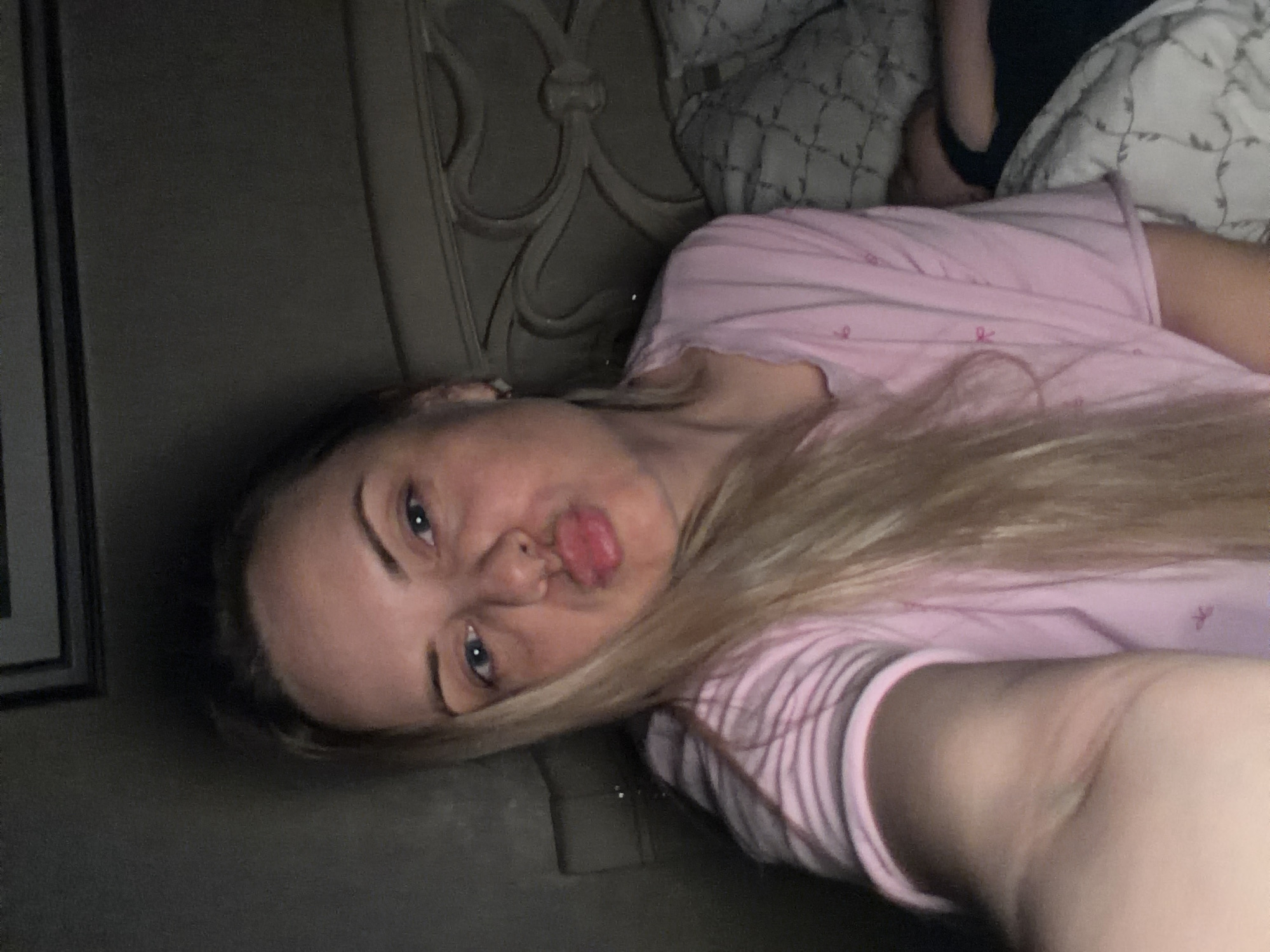 The image shows a woman with blonde hair lying on a bed. She is wearing a pink shirt. The headboard of the bed has a decorative design. A pillow with a patterned cover is visible behind her. A portion of a framed picture is seen on the left side of the image. It is not possible to determine the location of the image as Votkinsk, Russia from visual data alone.