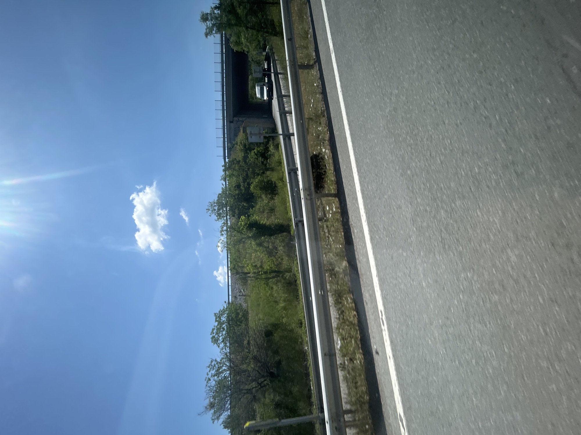 The image depicts a roadside scene under a clear blue sky with a few scattered white cumulus clouds. The sun is intensely bright in the upper right, casting significant lens flare. In the foreground, a paved asphalt road surface is visible, bordered on the right by a solid white line. Parallel to the road, a metal guardrail with two horizontal bars extends across the middle ground. Beyond the guardrail, a green, overgrown embankment rises, densely covered with various trees and bushes. In the mid-ground, a concrete overpass or bridge spans horizontally across the scene. Below the bridge, an underpass or tunnel section is visible, with dark graffiti on its internal concrete walls. A white van or small truck is partially visible exiting or entering the underpass on the far side, accompanied by what appears to be another vehicle or trailer behind it. The overall scene is brightly illuminated by direct sunlight. The location is in the city of Unknown, Unknown.