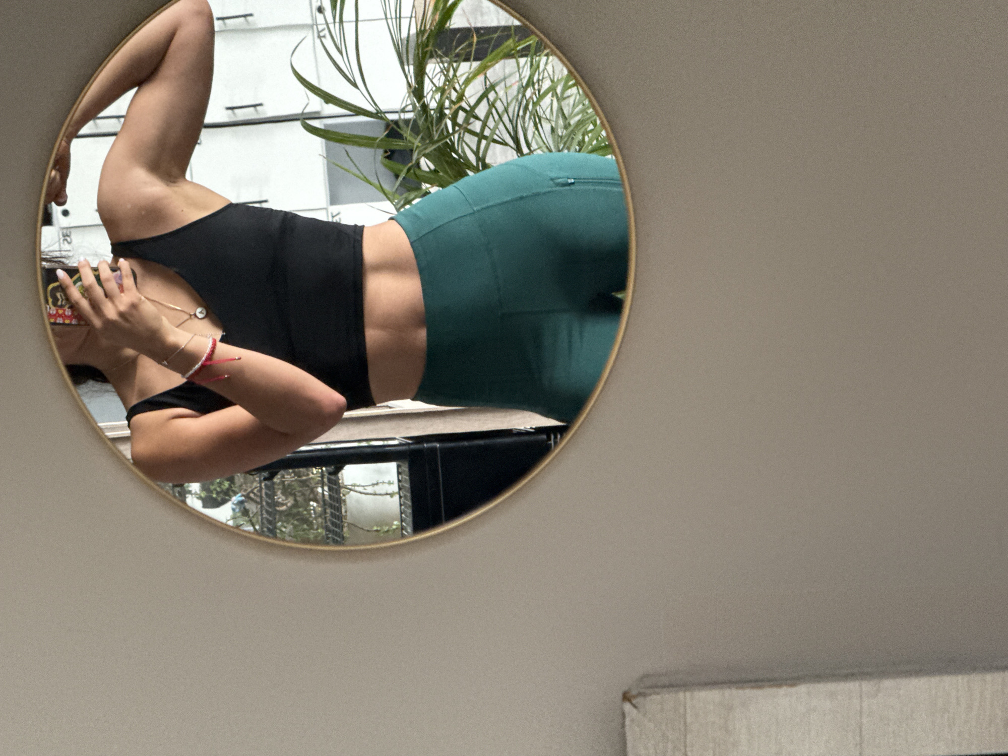 A woman in a black tank top and teal leggings is reflected in a round mirror. She is holding a phone in her left hand, and her right arm is raised. The background visible through the mirror includes a building and a plant. The mirror is mounted on a wall. The location is Puebla, Mexico.