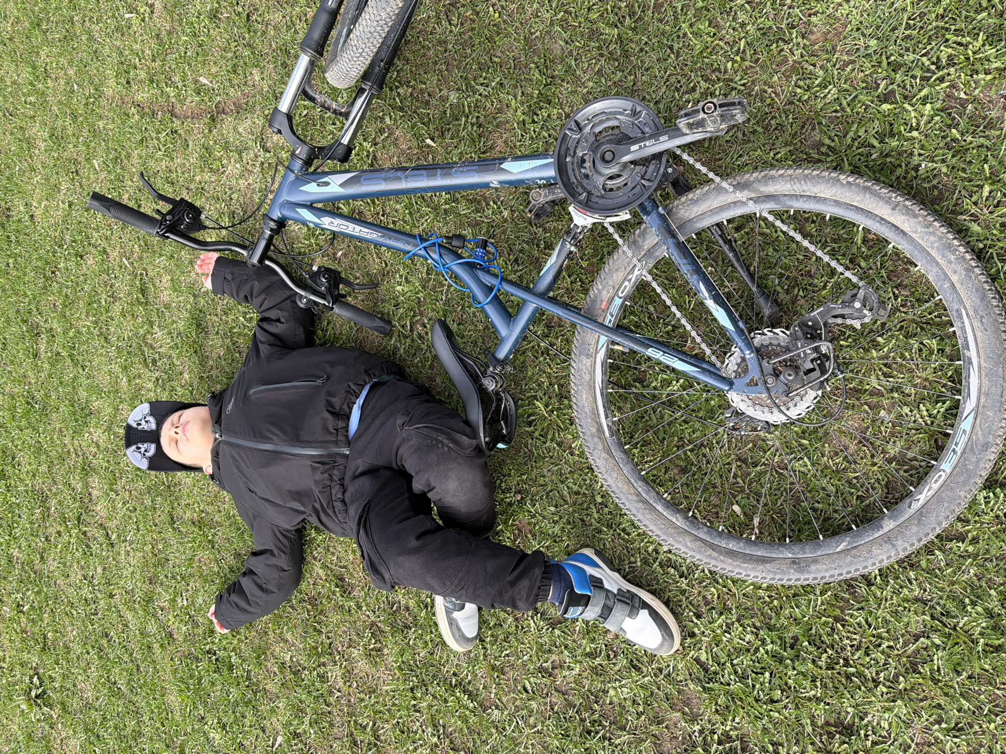 An overhead shot captures a child lying on their back in a grassy area next to an overturned bicycle. The setting is outdoors in bright daylight, likely in Votkinsk, Russia.
The child is dressed in a black zip-up jacket, black pants, and athletic shoes—one with grey and white, the other with blue and white accents. They wear a black baseball cap adorned with two white skull patches. The child's eyes are closed, and their body posture appears relaxed, suggesting they might be resting, playing, or possibly pretending to be asleep or unconscious, rather than in distress. Their arms are slightly outstretched, with one hand near the bike's handlebars.
Beside the child lies a blue mountain bike, identified by the visible text "STELS" and "NAVIGATOR" on its frame. The bike is on its left side, displaying its knobby tires, chain, gears, and pedals. The handlebars feature black grips and brake levers. A blue coiled cable lock is visible wrapped around the bike's frame. On the tire or rim, the number "26" is clearly visible, likely indicating the wheel size, along with partial text that appears to be "XDS."
The ground is covered in green grass, which shows some signs of wear in places. The overall impression of the scene is one of leisure or playful rest, with no immediate indications of an accident or injury.