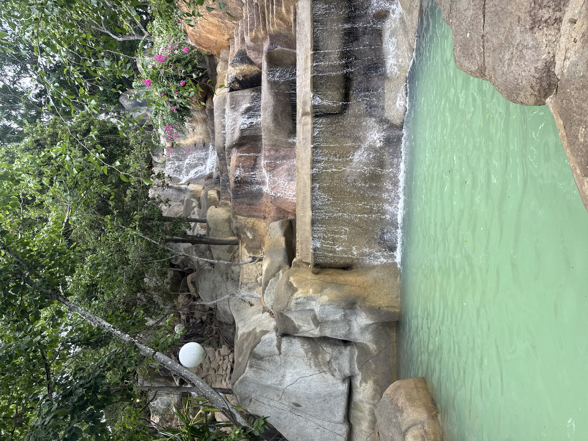 The image depicts a multi-tiered waterfall flowing into a turquoise pool. The waterfall is constructed with natural-looking rocks and concrete, with several cascades of water. Lush green trees and foliage surround the waterfall and pool area. In the background, there are also some pink flowers blooming on a bush. The scene appears to be an artificial water feature, likely part of a resort or park. Given the context of Nha Trang, Vietnam, this could be a feature within Vinpearl Land or a similar attraction. There are no people visible in the image. The lighting suggests it is daytime, with overcast skies contributing to a soft, diffused light. The water is clear and still in the pool, with ripples created by the falling water. The overall impression is one of a serene and tropical environment.