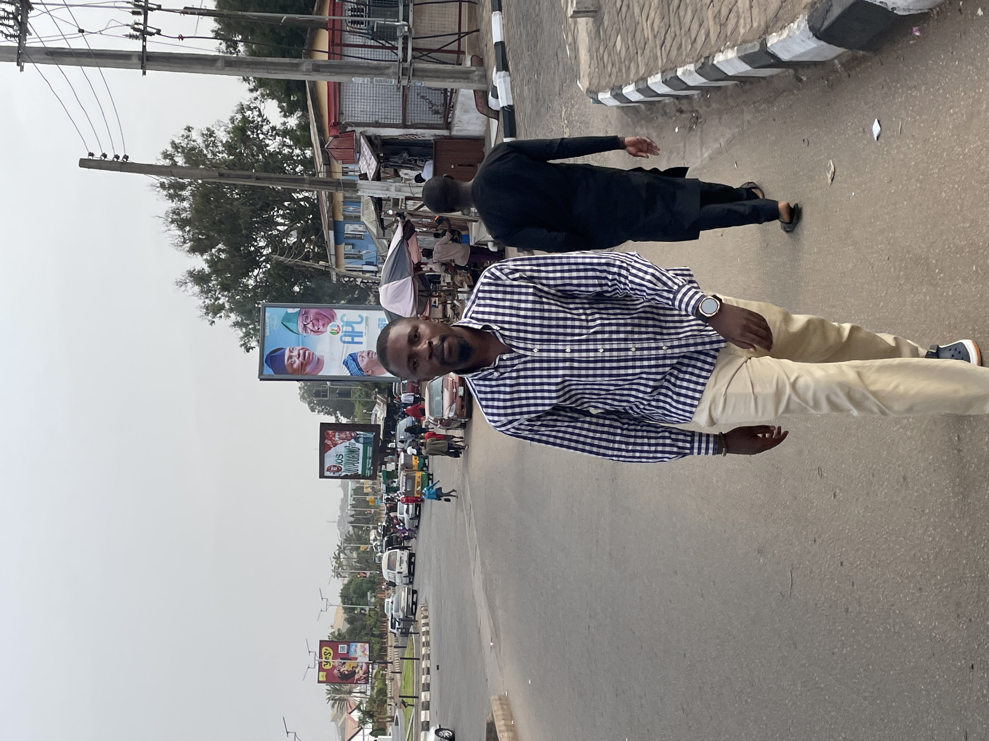 The image shows a street scene in Jos, Nigeria. A man in a checkered shirt and khaki pants is standing in the foreground, facing the camera. Another man in a black robe is walking away from the camera in the background. Cars and billboards line the street. Overhead power lines are visible.