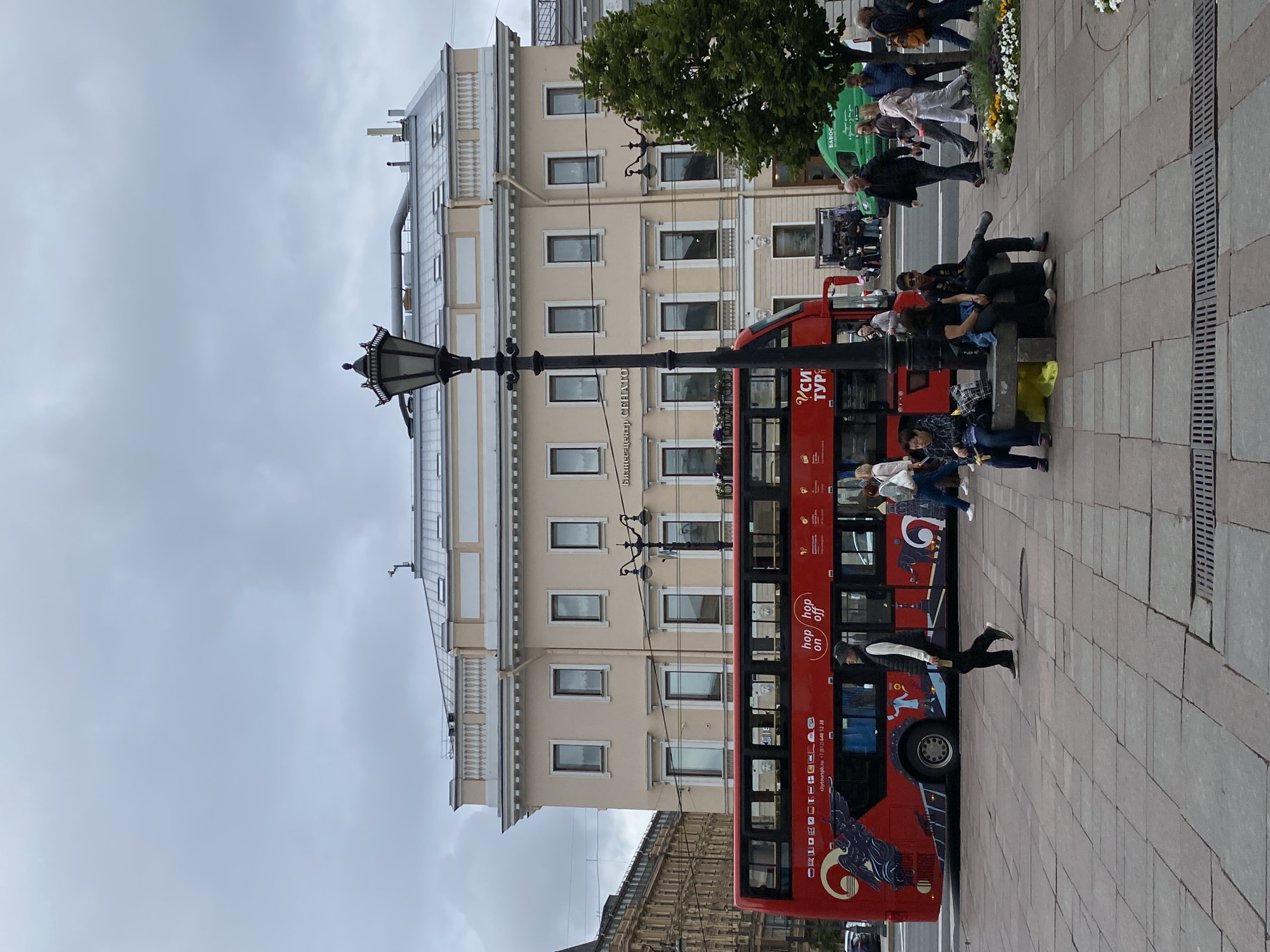 The image shows a street scene in Saint Petersburg, Russia, likely near a historic district given the architecture. A bright red double-decker bus, labeled "hop on hop off," is prominently featured on the left side of the frame. The bus appears to be a tour bus, as suggested by its design and advertisements. People are seen walking on the sidewalk and some are sitting on a bench in the foreground. The background features a large, light-colored building with numerous windows and decorative elements, typical of St. Petersburg's classical architecture. A lamppost stands in the middle ground. The sky is overcast, suggesting a cloudy day. There is also a green van visible on the right side of the street. The ground is paved with large stone slabs.