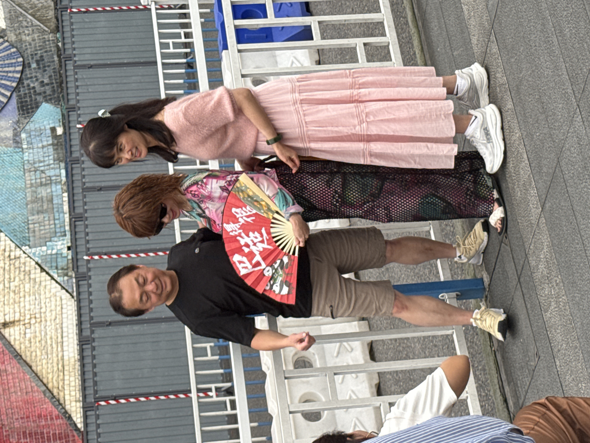 In Yuzhong District, China, a group of people stands alongside a metal railing. A woman in a pink sweater and layered skirt stands at the front, glancing downwards. Behind her, a woman with auburn hair wears a patterned shirt and a black skirt with a mesh overlay. A man in a black shirt and khaki shorts holds a red folding fan with white lettering and an image. The group stands on a grey paved surface. Sections of colorful architecture and metal barriers are visible in the background. Another person wearing a striped blue shirt and brown pants stands near the bottom edge of the photo.