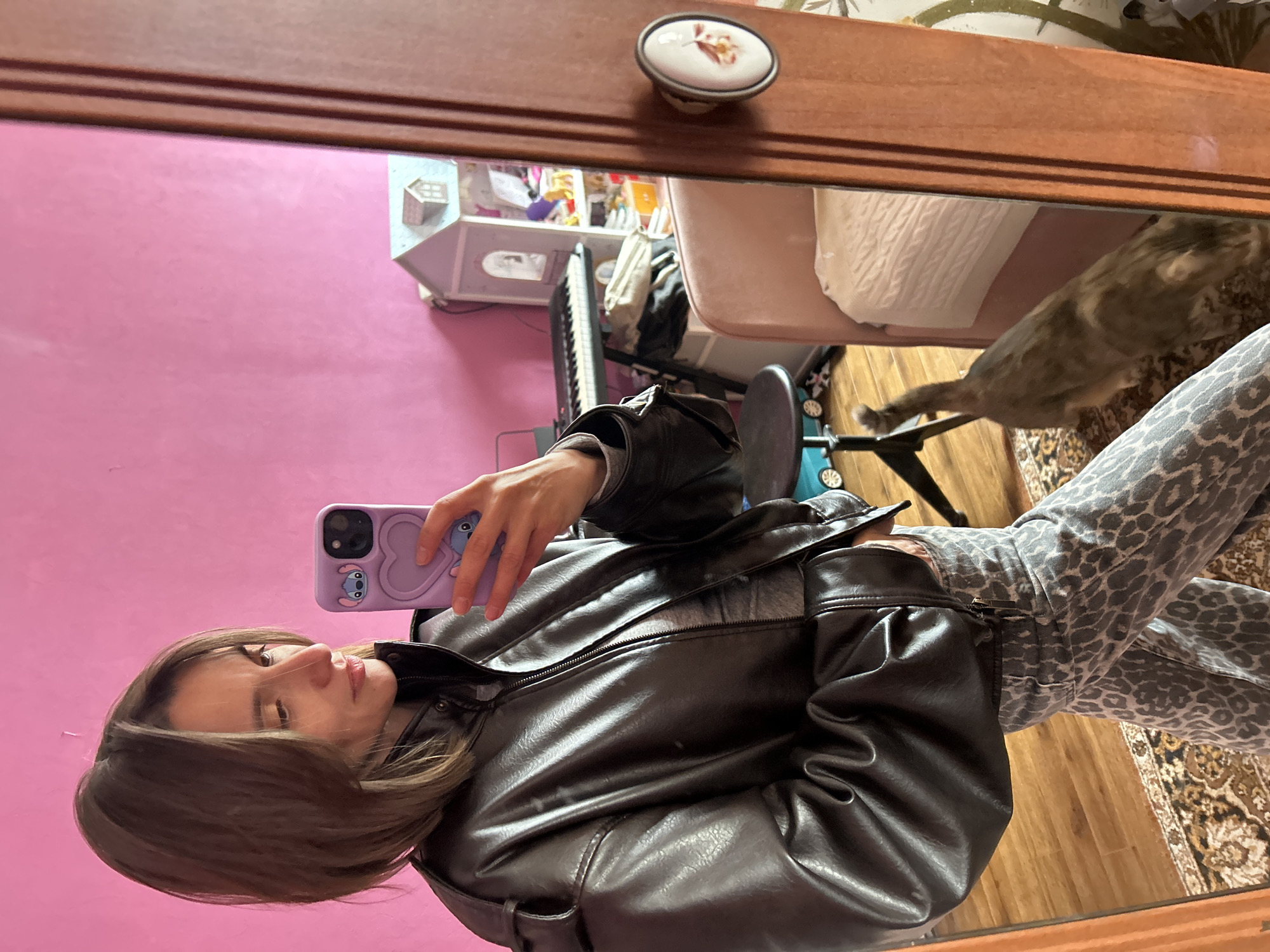 A person takes a selfie in a mirror. The person has brown hair, a leather jacket, and leopard print pants. The person holds a purple phone case with a blue cartoon character on it. The background shows a pink wall, a keyboard, a toy house, a chair, a bed with a white comforter, a cat, and a patterned rug. The location is an indoor space with personal belongings. It is impossible to determine if it is Moscow, Russia from the provided image.