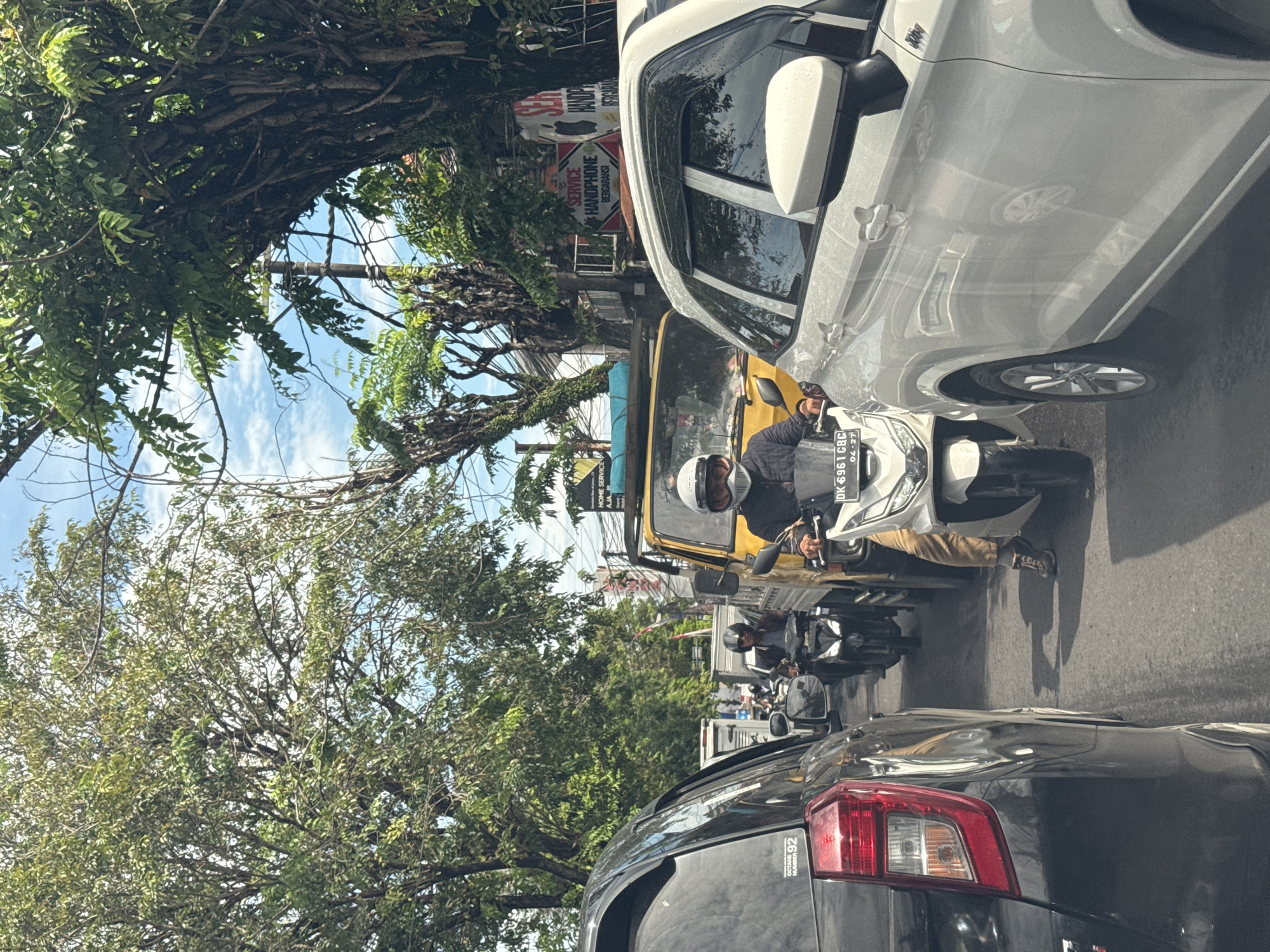 This media file captures a street scene in North Kuta, Indonesia, during the daytime. The perspective appears to be from inside a vehicle, looking out at traffic on a sunny day with a partly cloudy blue sky.
The main subjects include several vehicles and people. In the immediate foreground, the rear of a black SUV, displaying "OCTANE NUMBER 92" on its tailgate, is visible. To the right, a white car is prominent. The central focus is a person on a white scooter/motorcycle, wearing a white helmet and a dark jacket, seemingly paused in traffic. The scooter's license plate, "DK 6961 CBC," with "04-27" below it, confirms the Indonesian location, specifically Bali (DK code). Behind this motorcycle, a yellow vehicle and another motorcyclist wearing a dark helmet are visible, along with additional vehicles in the distance.
The activity taking place is typical urban traffic, with vehicles, especially motorcycles, navigating a busy road. Lush green trees with dense foliage and thick trunks line both sides of the street, suggesting a tropical environment.
Notable details include a building in the background featuring a red sign that reads "SERVICE HANDPHONE BERGARANSI" (Guaranteed Handphone Service), with "WANDRA" also partially visible. Shadows are cast on the road, indicating bright sunlight.