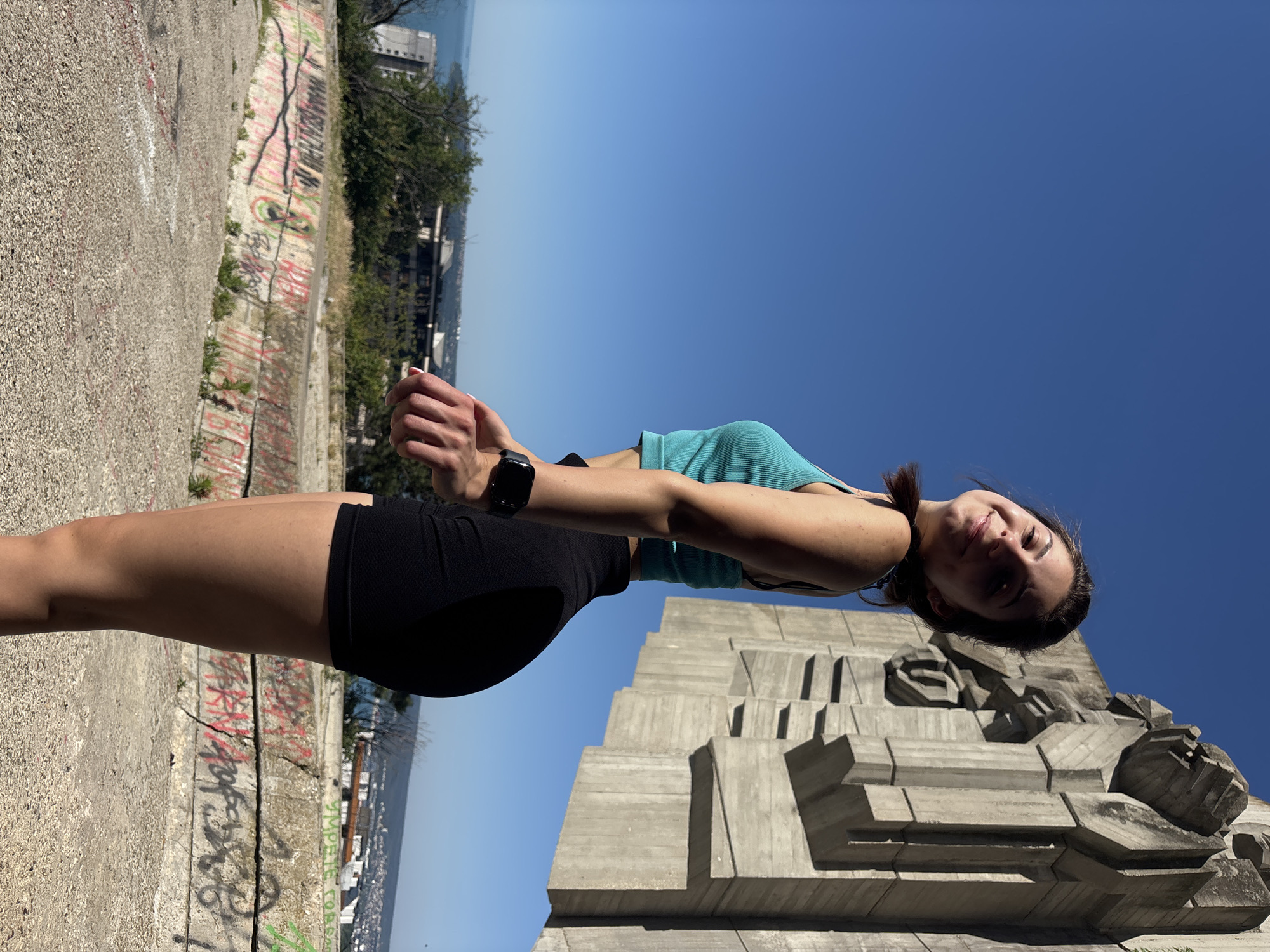 A young woman stands with her back to a large, abstract concrete monument against a clear blue sky. She is wearing a turquoise tank top and black shorts. Her left arm is extended and her hand is open, palm up, as if reaching out. She is smiling and looking towards the camera. In the background, a body of water can be seen, suggesting a coastal location. Below the monument and in the foreground, there are concrete structures covered in graffiti. The image was taken at the Monument to the Bulgarian-Soviet Friendship in Varna, Bulgaria.