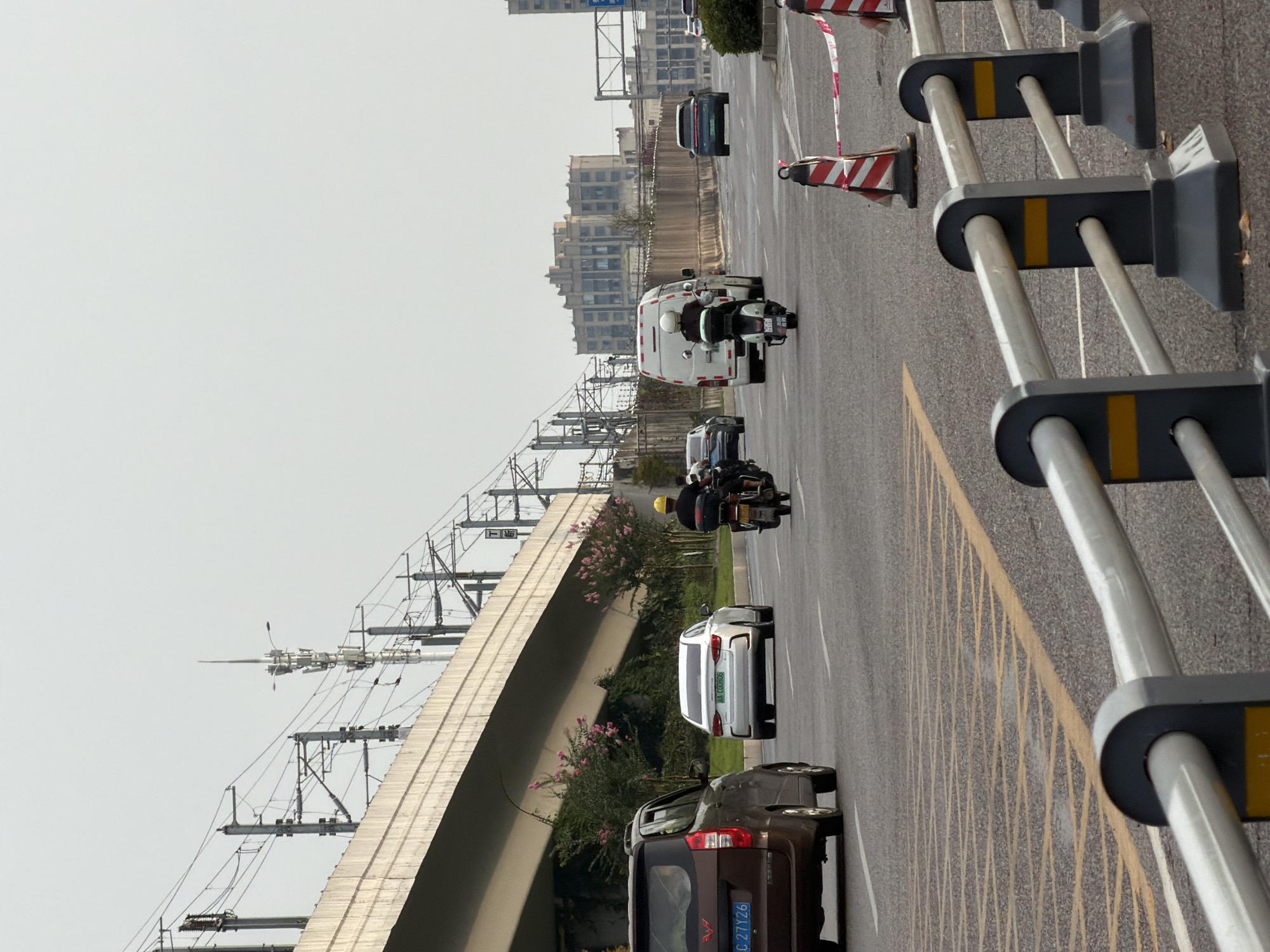 The image depicts a street scene in Jiangjin, China, characterized by elevated infrastructure and vehicular traffic. In the foreground, a metal guardrail runs horizontally across the lower portion of the frame, partially obscuring a road marked with yellow diagonal lines, suggesting a traffic calming or merging area. Beyond the guardrail, traffic moves along a multi-lane asphalt road.
Several vehicles are visible: a dark brown SUV is on the left, a white sedan is in the middle lane, and a white van and a scooter are further ahead in the right lanes. Two individuals are operating scooters, one with a yellow helmet and the other with a white helmet. The background features an elevated concrete structure, likely a highway or railway overpass, supported by concrete pillars. Overhead, complex electrical wiring and utility poles are suspended, indicating a significant transportation network. In the distance, multi-story buildings with an aged facade are visible under an overcast sky. The overall environment appears to be an urban or suburban thoroughfare.
