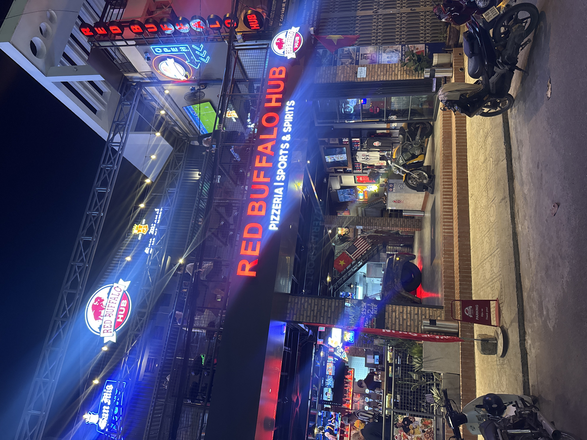 The image shows a street view at night of "RED BUFFALO HUB", a business located in Bac Nha Trang Ward, Vietnam. The establishment features prominent neon signage, with "RED BUFFALO HUB" illuminated in red letters above the entrance. The facade is a mix of brick and modern architectural elements, with multiple levels and balconies. There are several televisions visible, some displaying sports. A yellow motorcycle is parked inside the entrance, and a dark motorcycle is parked on the sidewalk in the foreground. A red flag and a "PARKING" sign are also present on the sidewalk. Several people are visible inside the establishment, including at a bar area. The street is paved, and there are overhead lights and strings of lights illuminating the scene. The overall atmosphere is that of a lively entertainment venue.