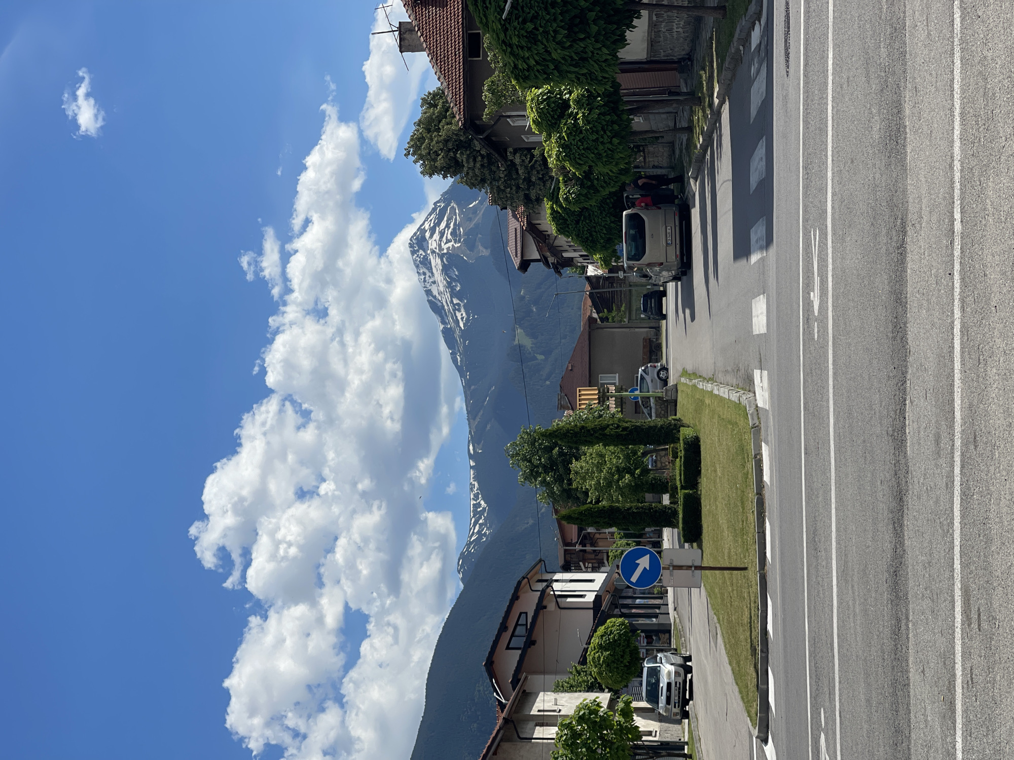 An eye-level, vertical, medium shot captures a street scene in a town at the base of snow-capped mountains under a bright blue sky with fluffy white clouds. In the foreground, a paved road with white lane markings and a pedestrian crossing occupies the lower half of the frame. On the left side of the road, a silver SUV is parked next to a sidewalk and a small grassy area with manicured bushes and a blue sign with a white arrow pointing to the right. Beyond the sidewalk are buildings with light-colored facades and dark trim, one of which has large windows. On the right side of the road, the sidewalk is lined with trees with rounded tops, and more buildings with reddish-brown tiled roofs and stone bases. A light-colored van is parked on this side of the street, with two individuals standing near its open rear door. In the background, a range of dark, tree-covered mountains rises, with their peaks dusted with snow and illuminated by the sun. The overall impression is of a picturesque mountain town on a clear, sunny day.