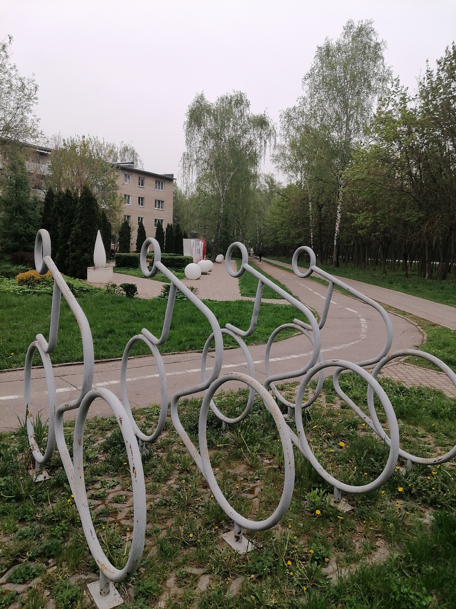 The image shows a public space in Uspenskoye, Russia. In the foreground, there are four metal bicycle racks shaped like bicycles, painted in light gray. The racks are positioned on a patch of grass and a paved surface with weeds and yellow flowers growing around them.
Behind the bike racks, there is a walking and bike path with a painted white line, indicating lanes. Further back, there is a lawn with sculptures and trees. A building with a light-colored facade and multiple windows is visible behind the trees. The sky is overcast, suggesting a cloudy day. A person is visible walking in the distance on the path.