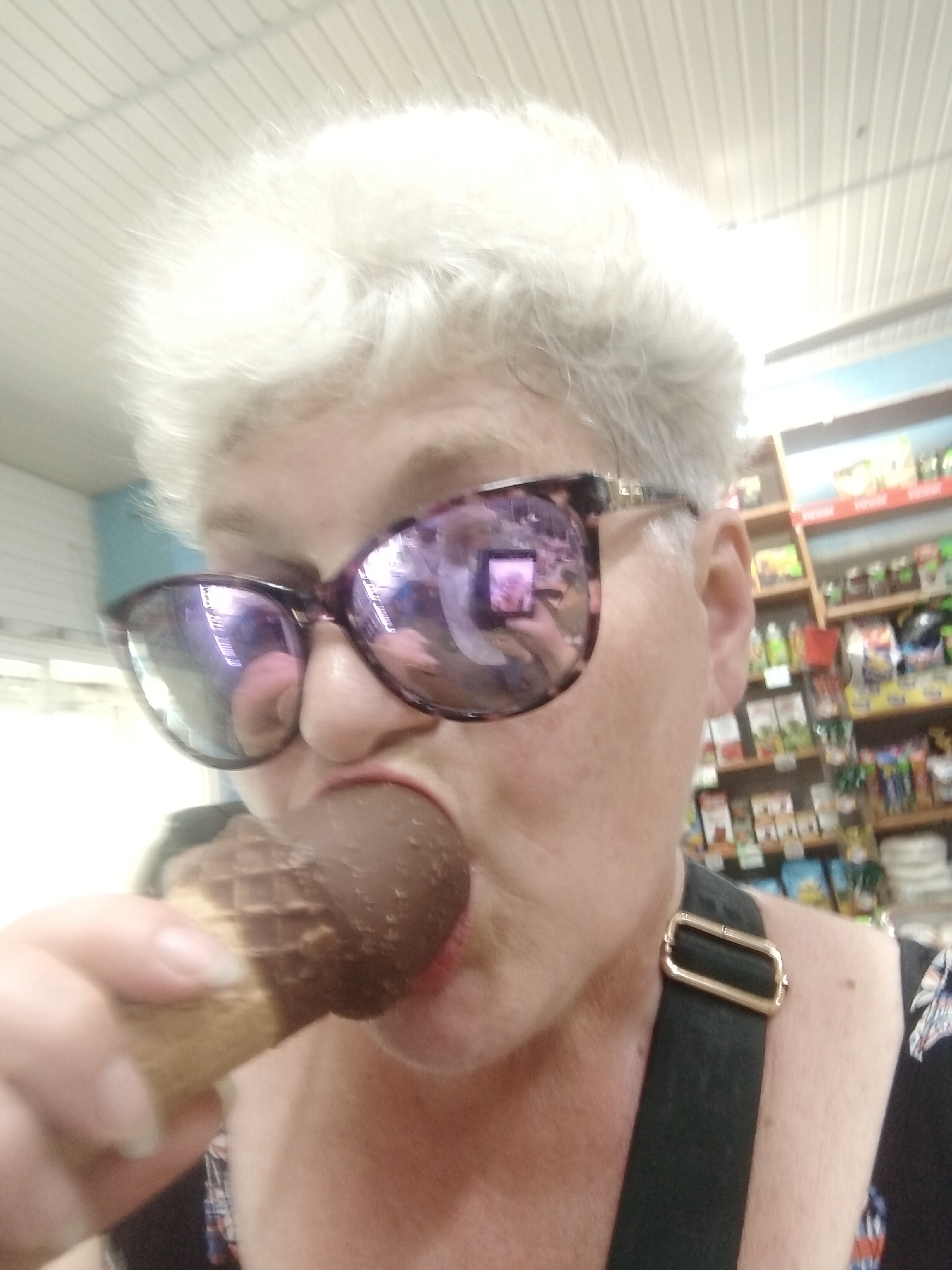 The media file is a close-up, selfie-style photograph of an older woman with short, white hair, wearing large, purple-tinted sunglasses. She is in the process of eating an ice cream cone that is covered in chocolate. Her mouth is open, and the ice cream cone is in her mouth, with the chocolate-covered portion visible.
The setting appears to be indoors, possibly a store or market. In the background, shelves are stocked with various items, including what seem to be food products or toiletries. The lighting suggests it is daytime. The woman's expression is difficult to discern due to the sunglasses, but the act of eating ice cream often implies enjoyment or a casual moment.
A black strap with a gold buckle is visible on her shoulder, suggesting she might be wearing a bag or an apron. The overall mood of the image is informal and personal. There is no visible text in the image. The location context is Horishni Plavni, Ukraine.