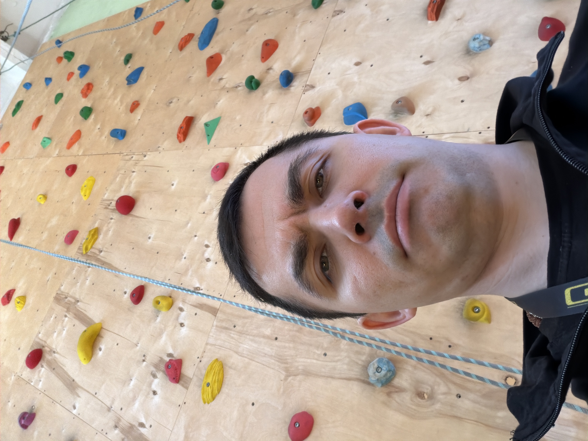 A man is taking a selfie while rock climbing. He is wearing a harness and standing in front of a climbing wall with multi-colored holds. The background appears to be a gym setting. There is no indication that the photo was taken in Izhevsk, Russia.