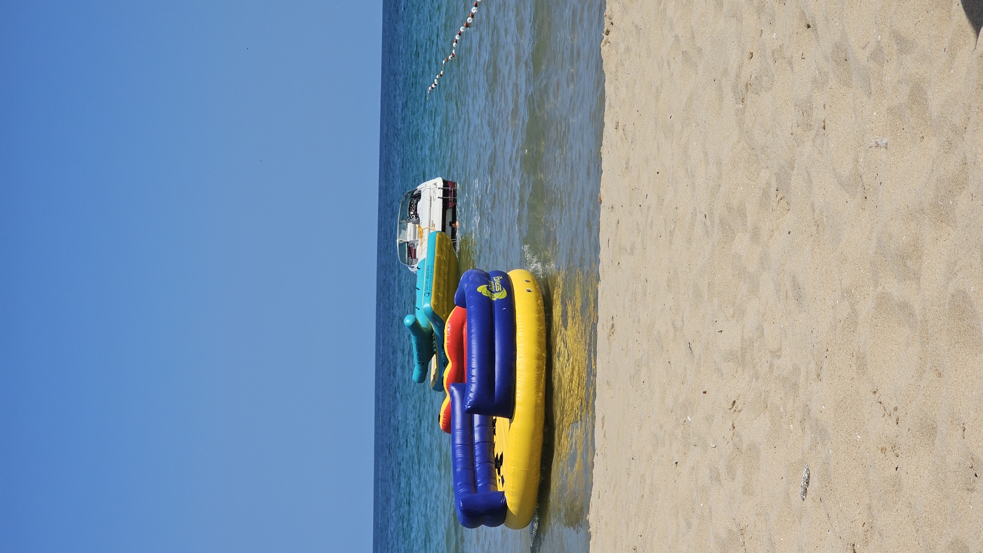 This is a vertical, eye-level, medium shot of a sandy beach with the sea and sky in the background. In the foreground, a tan sandy beach slopes gently towards the water. The sea is a clear blue with small waves lapping at the shore. In the water, there are two inflatable water toys and a small white boat. The closest inflatable toy is yellow with blue sides and red accents. The second inflatable toy is blue-green and shaped like a sea creature. The white boat is docked behind the inflatable toys. The sky is clear and bright blue. There is a rope with buoys visible in the water in the distance. There are no people visible in the image. The scene is consistent with the beaches found along the coast of Varna, Bulgaria.