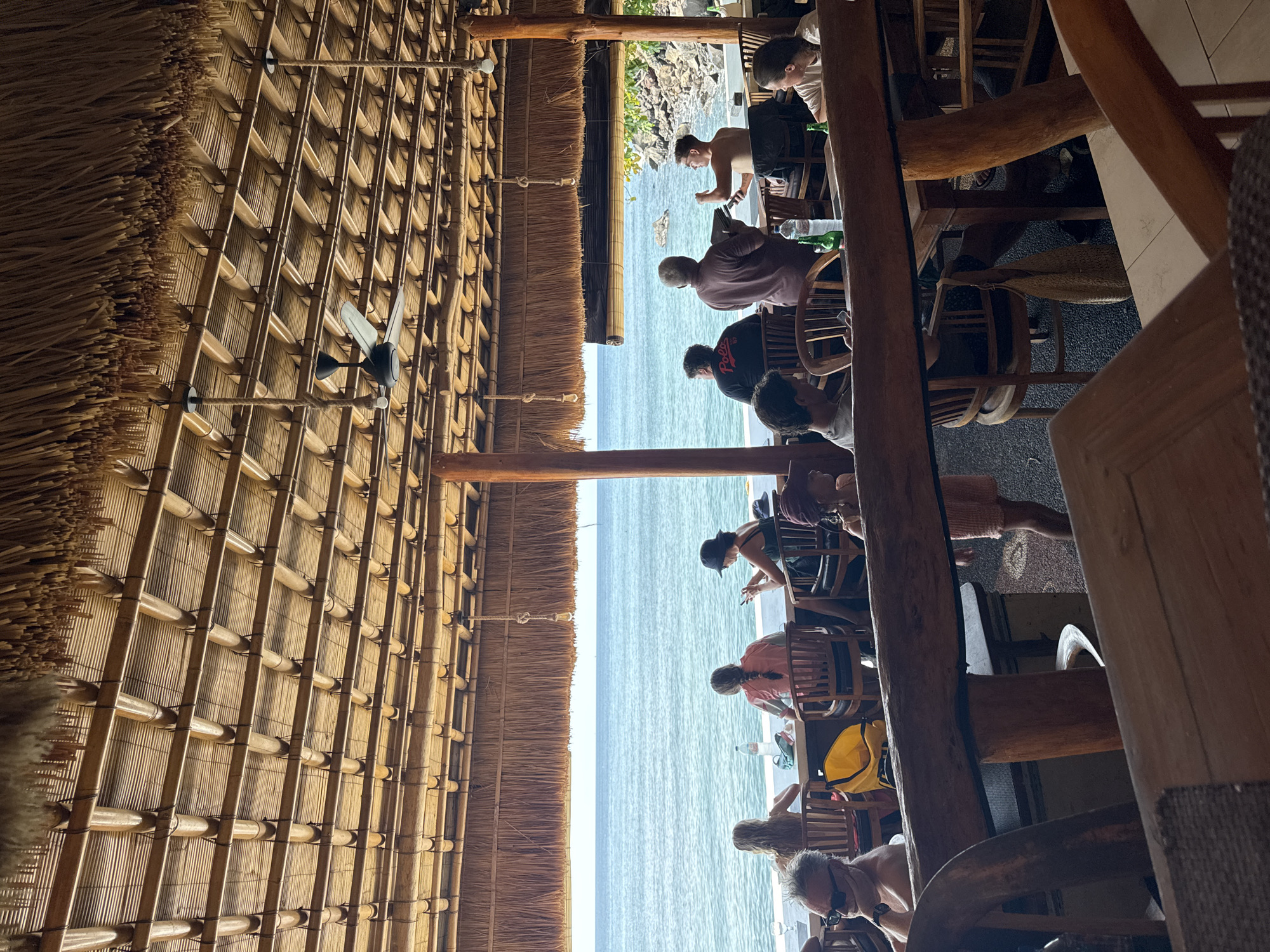 This image depicts a waterfront restaurant in Abang, Indonesia, featuring a thatched roof ceiling supported by bamboo beams. The restaurant overlooks the ocean, with a rocky shoreline visible to the right. Several patrons are seated at tables, some facing the water. In the foreground, wooden furniture and railing elements are prominent. A ceiling fan is suspended from the bamboo structure. The overall ambiance suggests a relaxed, tropical dining setting.