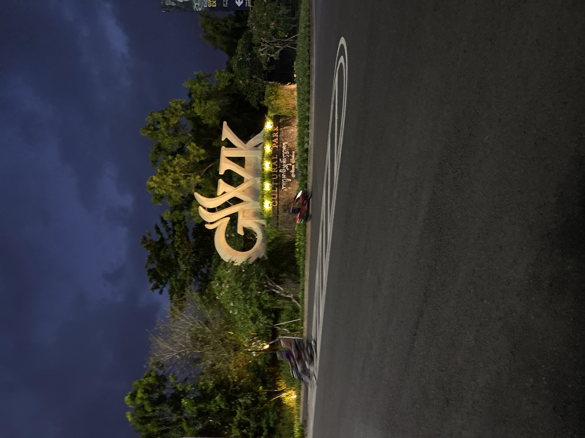 The image, taken at night in Kuta Selatan, Indonesia, depicts a large, illuminated sign that reads "GWK CULTURAL PARK" in English and Balinese script. The sign is predominantly white and stands out against a dark, cloudy sky. Lush green trees and foliage surround the sign, with some trees appearing bare or sparsely leafed.
In the foreground, a dark asphalt road is visible, marked with white lines, suggesting it's a pathway or entrance. A few objects are on the side of the road near the sign. One is a red and black motorcycle, and further down, a dark bicycle or scooter with a person seated on it is visible. The lighting suggests it is nighttime, and the cloudy sky indicates overcast weather. The overall mood is quiet and serene, with the illuminated sign being the focal point. There is also a vertical banner or sign visible in the top left corner with some text, but it is largely out of focus and illegible.