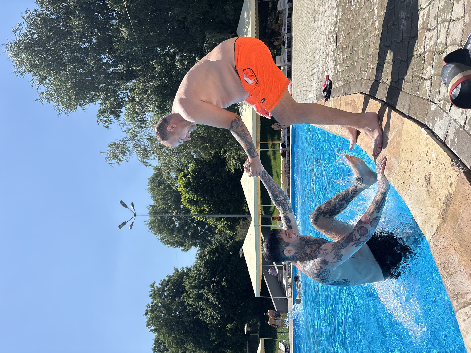 A man with extensive tattoos on his arms and torso is in a swimming pool, with his legs kicking and creating a splash. Another man, wearing orange swim trunks, is standing on the pool deck, reaching down to help pull the tattooed man out of the water. Both men appear to be smiling and engaged in a playful interaction. The pool area is outdoors, with lounge chairs and umbrellas visible in the background. Trees and a clear blue sky suggest a sunny day. This scene is likely at a public swimming pool in Almaty, Kazakhstan, possibly during the summer months.