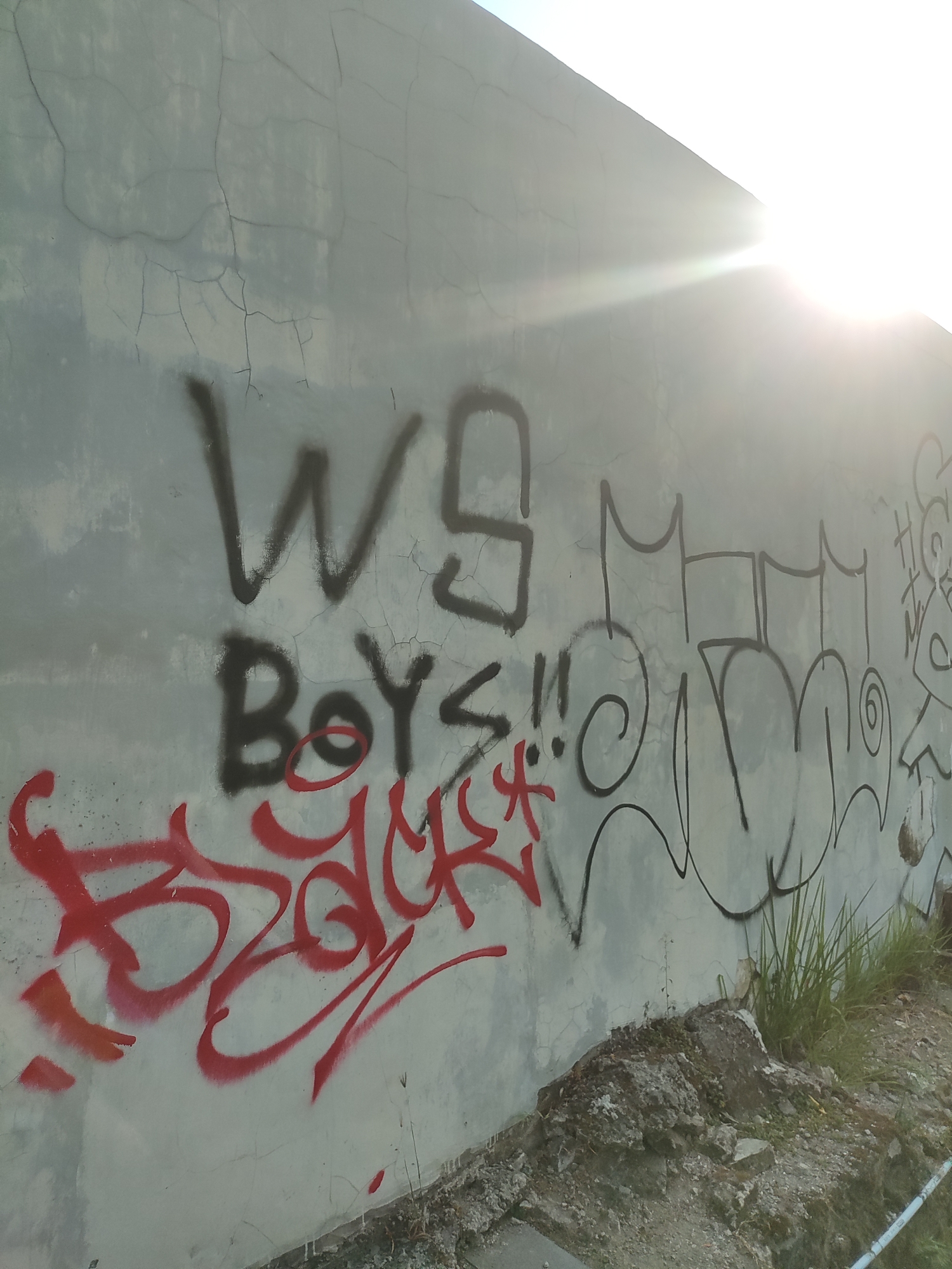 A slightly cracked, light gray concrete wall in Cirebon, Indonesia, is covered with graffiti. On the left side of the wall, the words "WS BOYS!!" are sprayed in black. Below "BOYS!!", red graffiti spray paint forms a swirling design that appears to spell out "BACKER". To the right of "WS BOYS!!", black graffiti spray paint forms stylized letters and a cartoonish face with an outline of a circle around it. The sun is shining brightly from the upper right corner of the image, creating lens flare and washing out part of the wall. The ground in front of the wall is covered in dirt, small rocks, and some sparse green grass.