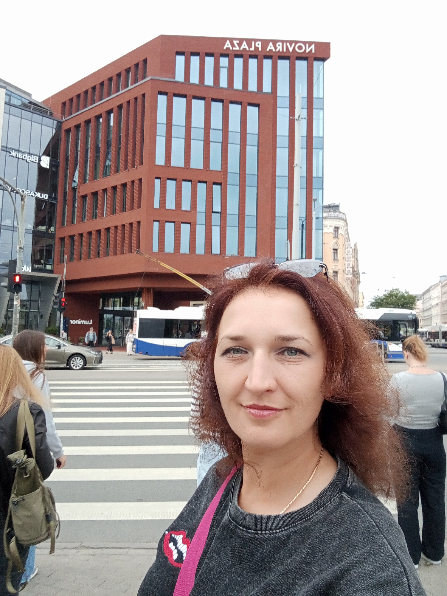 A mid-shot, outdoor, selfie-style photo captures a woman with reddish-brown hair, wearing a dark grey t-shirt and a pink strap across her shoulder. She is standing on a pedestrian crossing in front of a modern, red brick building. The building has numerous rectangular windows and a sign at the top that reads "ASADA ARIYON" in white letters. To the left of the red brick building is a glass-fronted building with the word "Bidpank" visible. A silver car is stopped at the pedestrian crossing, and a blue and white bus is further down the street. The sky is overcast. The scene appears to be a street in Riga, Latvia.