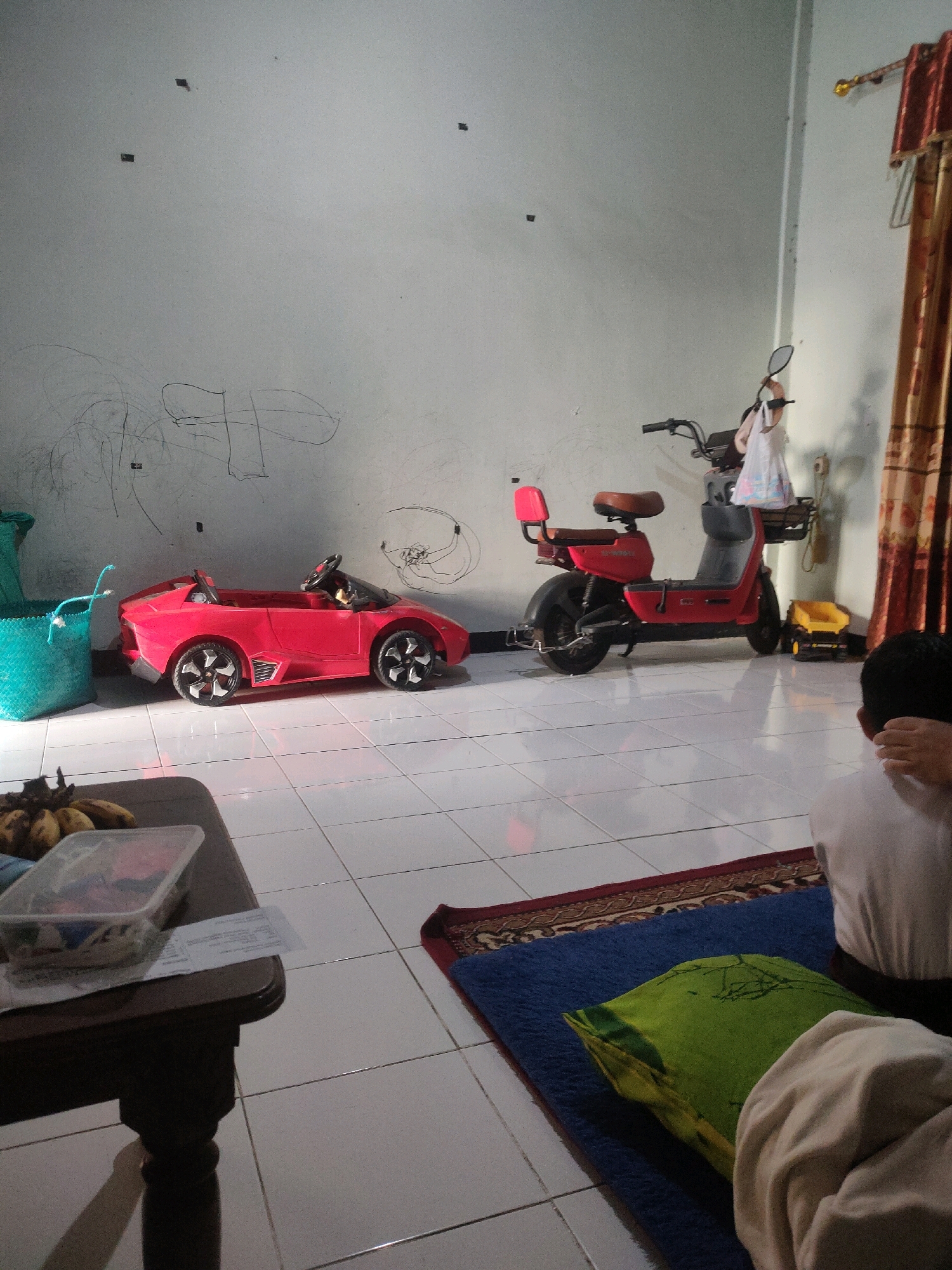 In a room with white tiled flooring and a white painted wall, a red toy sports car and a red toy scooter are parked side-by-side. The toy car is positioned to the left of the scooter. The wall behind the toys has children's scribbles on it. To the left of the toy car, a turquoise woven basket is visible. In the foreground, a dark wooden table holds a clear plastic container with bananas and other items. To the right, a person is sitting on a blue mat with a patterned rug underneath, and a green pillow is beside them. In the background, brown curtains hang on the right side of the wall, and a small yellow toy truck is on the floor next to the scooter. The scene depicts a domestic environment, likely a living room or playroom, in Tebuwung, Indonesia.