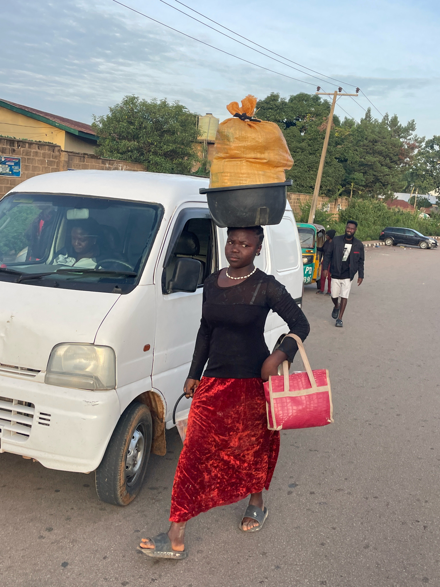A young woman carries a large, yellow, netted bag balanced on a black bowl on her head. She is wearing a black, long-sleeved shirt with sheer paneling on the upper chest and arms, a red velvet skirt, and dark sandals. She holds a pink textured tote bag in her right hand. The woman stands on a paved road next to a white van. In the background, a man in a black jacket and white shorts walks away from the camera, and other vehicles, including a small green and yellow bus, are visible. The environment suggests an urban setting, possibly in the city of Jos, Nigeria, with buildings and trees in the distance. The sky is overcast.