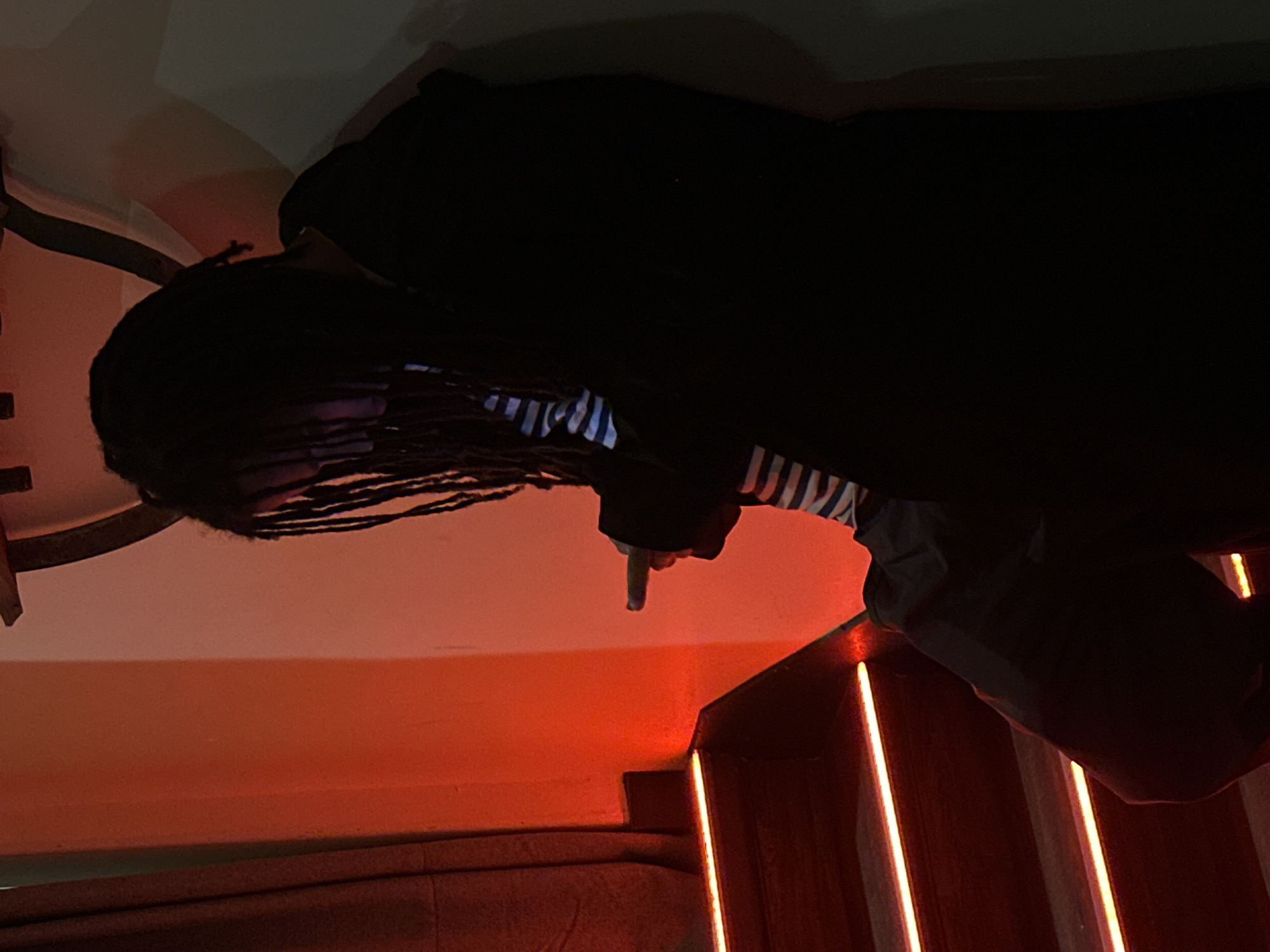 A person with dreadlocks, wearing a black jacket over a striped shirt, stands on a staircase illuminated by orange lights. The location is unknown.