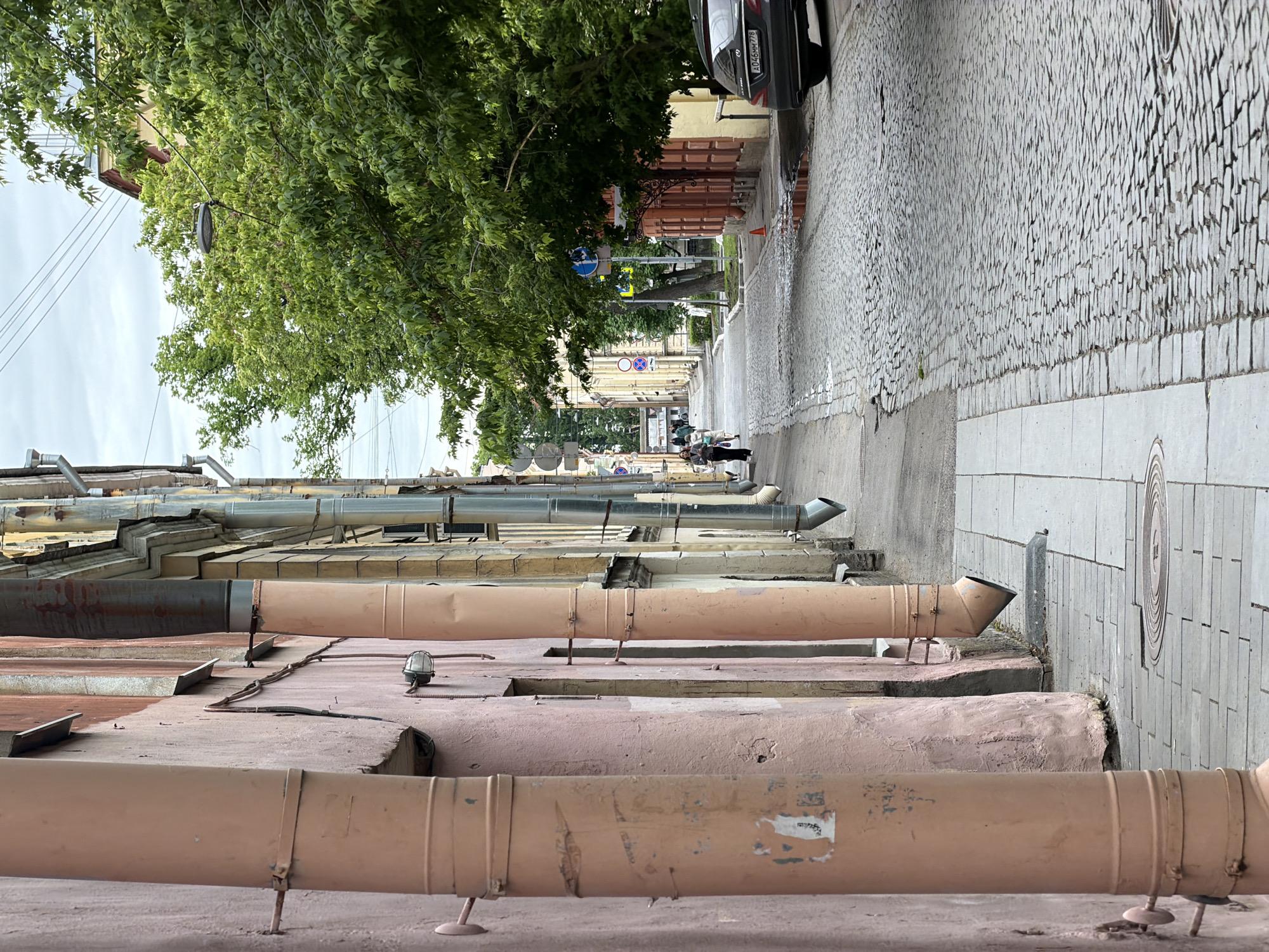 This image depicts a street in Saint Petersburg, Russia. The view is from the side of a building, focusing on the numerous exterior pipes and architectural features. In the background, the street stretches into the distance with a cobblestone road and sidewalks. A dark-colored car is parked on the right side of the street, partially visible. The presence of green trees and various street signs suggests an urban, residential area. Several people are walking in the distance. The overall tone is muted, typical of a European city street scene.