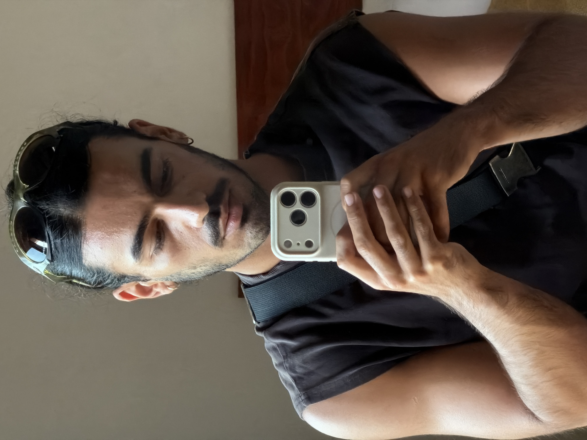 The image shows a man taking a mirror selfie, oriented 90 degrees counter-clockwise. The man, who appears to be of South Asian or Middle Eastern descent, has dark hair, a mustache, and a short beard. His eyes are closed or heavily lidded, conveying a relaxed or introspective expression. He is wearing a dark, sleeveless top or t-shirt. A pair of dark-lensed sunglasses is pushed up onto his head.
He holds a light-colored smartphone with a prominent triple-lens camera system in his right hand, reflecting his image. His arm is visible, showing fine body hair. A black strap with a silver metal buckle crosses his torso, possibly part of a bag or clothing accessory. The background suggests an indoor setting with light-colored walls and a glimpse of dark wooden furniture. The lighting indicates either daytime or a well-lit interior. No discernible text is visible on any objects in the frame. The provided context states the location is North Kuta, Indonesia, though this is not visually evident in the image.