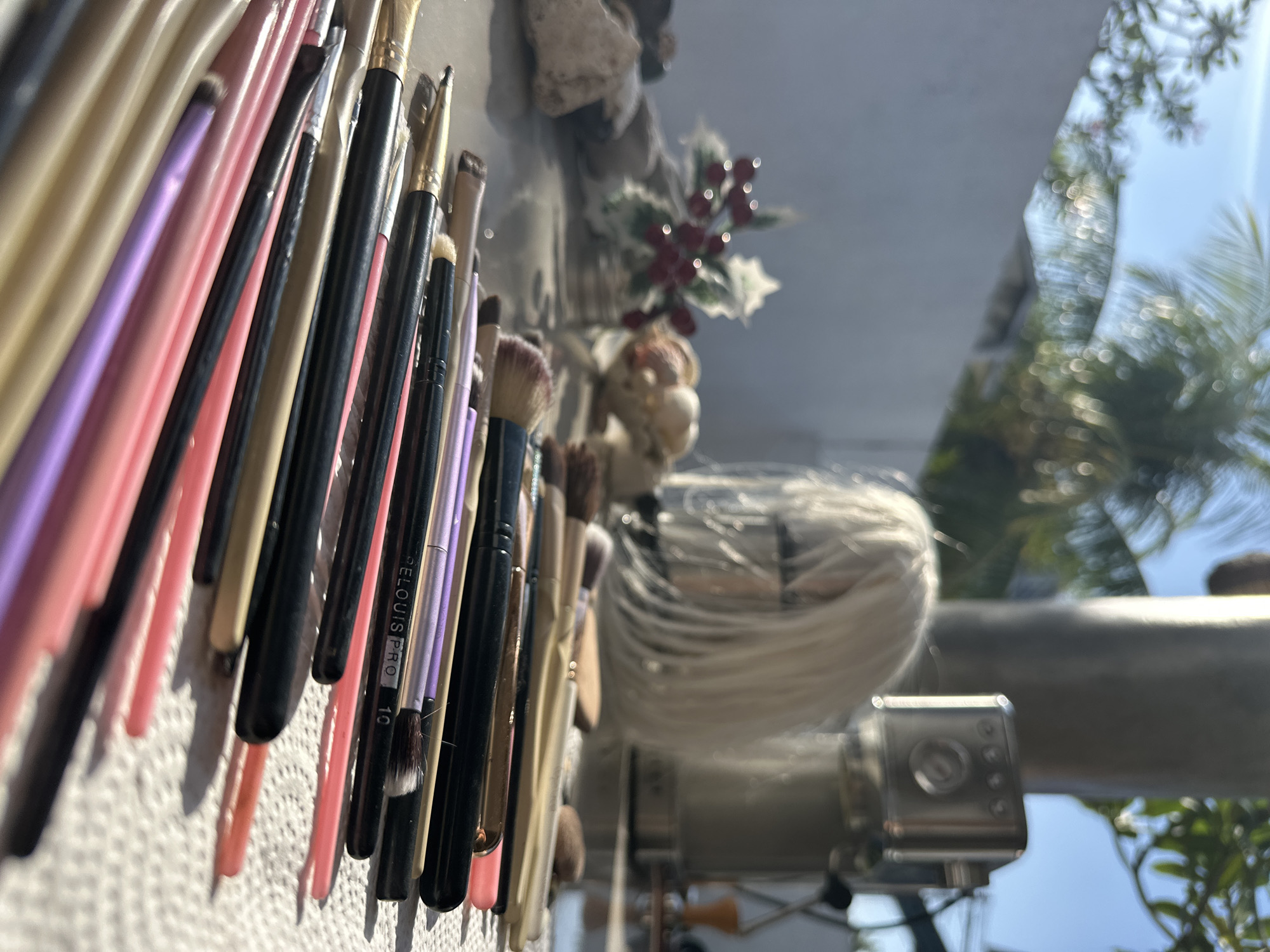 The image depicts a collection of makeup brushes arranged on a white, textured surface, possibly paper towels, outdoors. The brushes have handles in various colors including black, pink, purple, and gold, and their bristles are visible. In the background, out of focus, are elements of an outdoor setting: palm fronds, a bright blue sky, a white wall, and a metal object that could be a coffee machine. A wig on a mannequin head is also visible in the background, as is a decorative sprig of holly with red berries. The lighting suggests a sunny day. The scene is likely set in an outdoor area of a residence or studio in North Kuta, Indonesia, given the presence of tropical foliage.