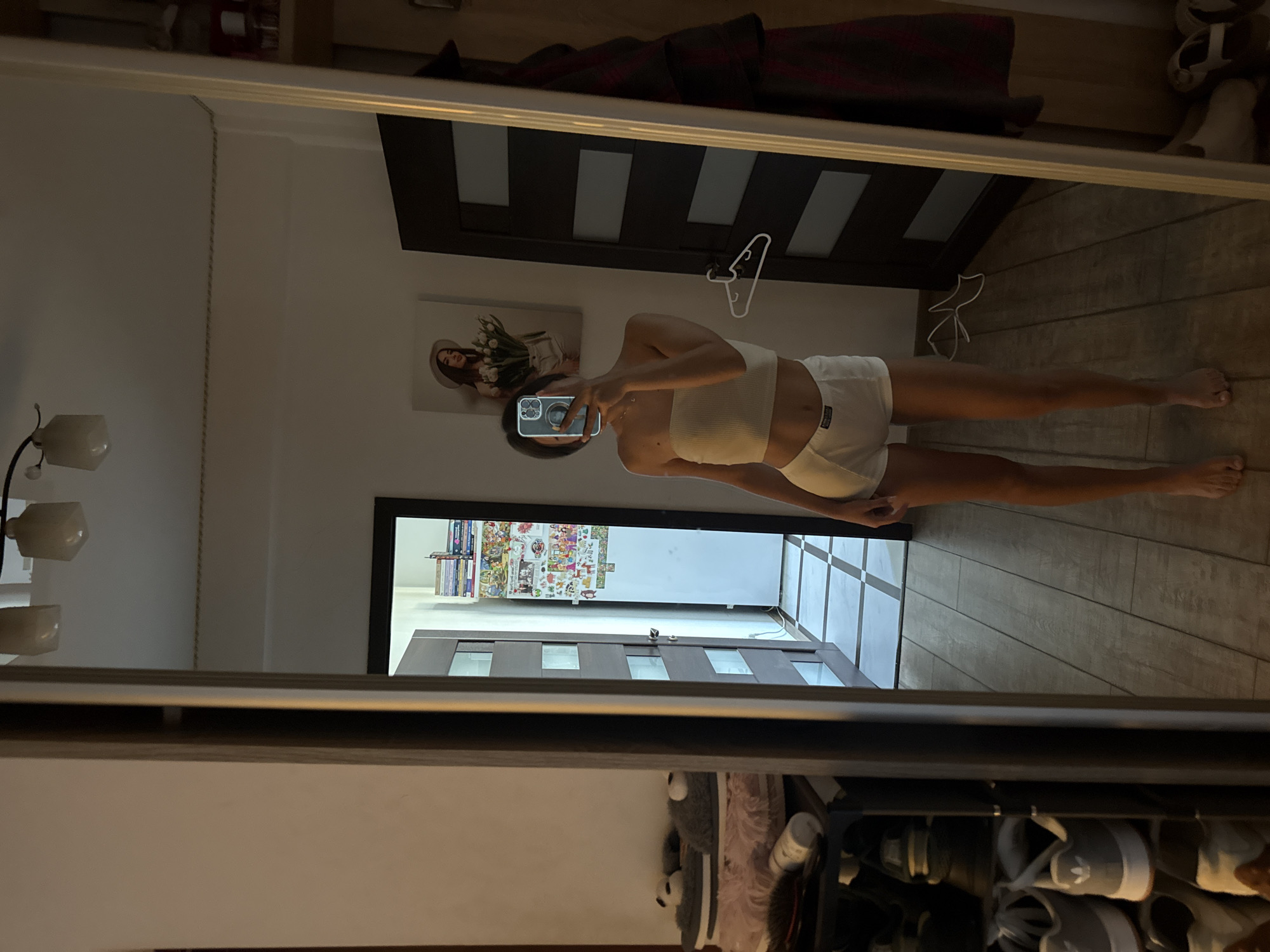 The image depicts a young woman standing in a well-lit room, taking a mirror selfie. She is wearing a white bandeau top and white boxer shorts. The room appears to be a bedroom or dressing area, with a large mirror dominating the right side of the frame.
The woman is positioned in front of the mirror, her right arm raised to hold a smartphone, obscuring her face. Her left arm is slightly bent, resting at her side. She is facing the mirror, and the reflection shows her full body.
To the left of the woman, a doorway opens into another room, revealing a shoe rack filled with various shoes. Further into this room, a white refrigerator is visible, covered in colorful stickers and what appears to be a stack of books.
On the wall behind the woman, to her left, a framed picture of a woman in a hat holding a bouquet of tulips hangs. A white clothes hanger is also visible on the door behind her. The floor is tiled with a pattern of light and dark squares in the background and wooden planks in the foreground.
The lighting in the room is soft, creating a warm ambiance. The overall impression is one of a casual, personal space. The city of Chișinău, Moldova, is not directly identifiable from the visual cues in the image.