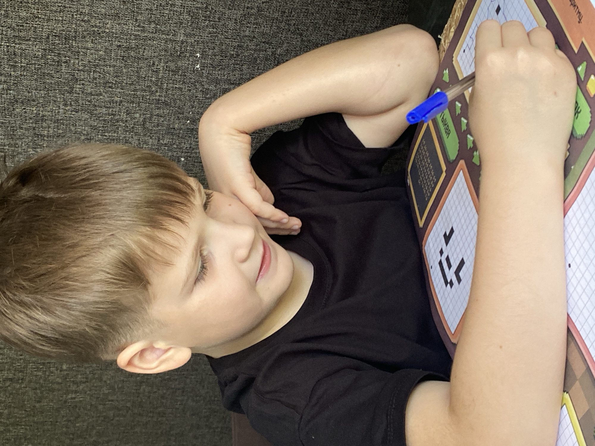 A young boy, wearing a black t-shirt, is sitting at a table and concentrating on a crossword puzzle. He is leaning his chin on his left hand and holding a blue pen in his right hand, poised to write in the puzzle. The crossword puzzle features pixelated graphics. The background is a textured, dark gray fabric, possibly a sofa or chair. The lighting suggests an indoor setting, possibly a home. The image was taken in the city of Chaikovsky, Russia.