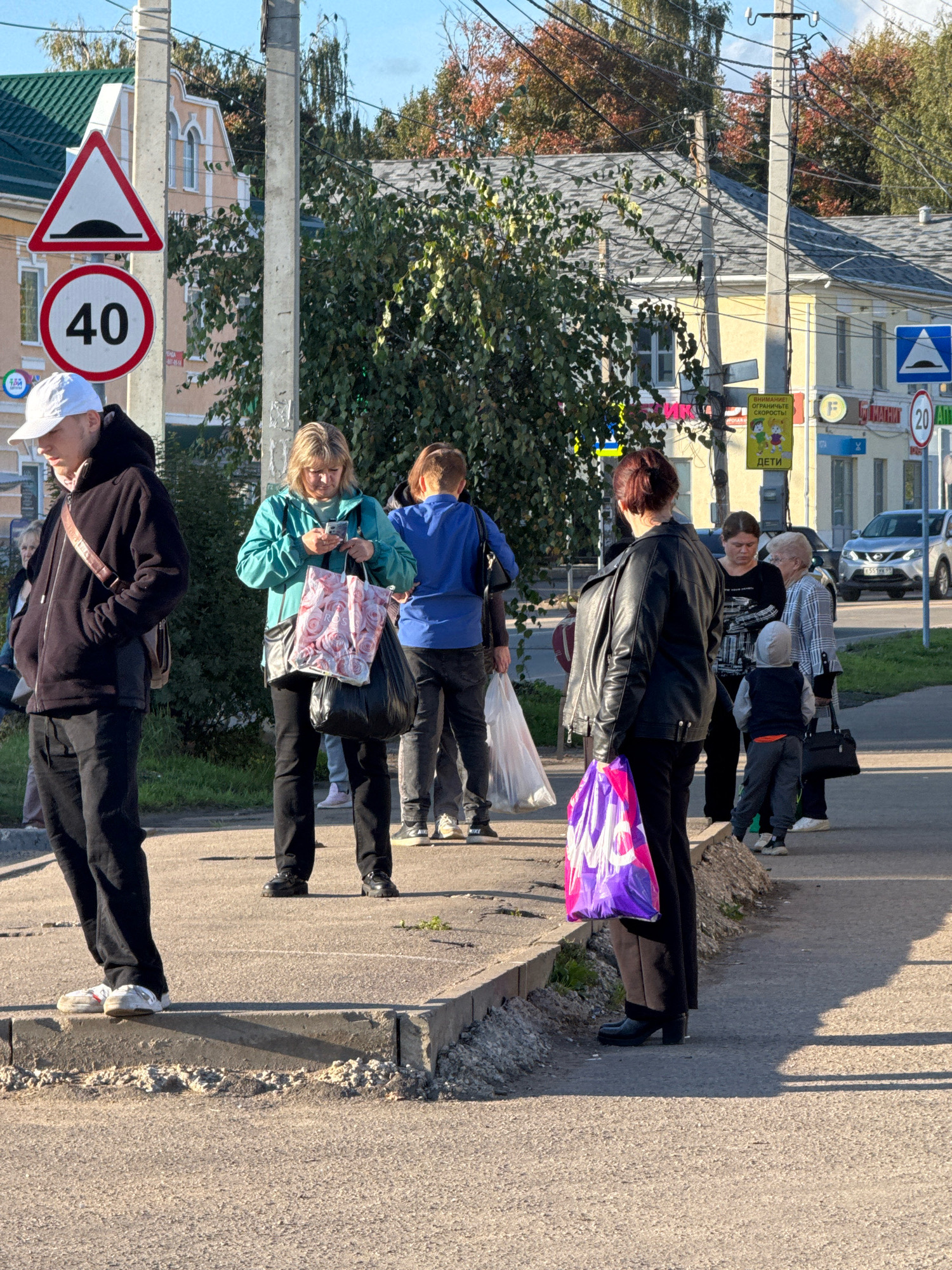 A group of people are standing on a sidewalk next to a street in Torzhok, Russia. In the foreground, a young man in a black hoodie and pants is facing left, looking down. To his right, a woman in a turquoise jacket is looking at her phone while holding two shopping bags. Behind them, a person in a blue shirt is standing, and further down the sidewalk, a woman in a black leather jacket is facing away from the camera, holding a purple and pink shopping bag. Other individuals are visible in the background, including a child in a grey hoodie. Traffic signs, including a speed limit sign indicating 40 and a warning sign for a speed bump, are posted on poles. Buildings, trees with autumn foliage, and power lines are also present in the environment. A car is parked on the street in the background.