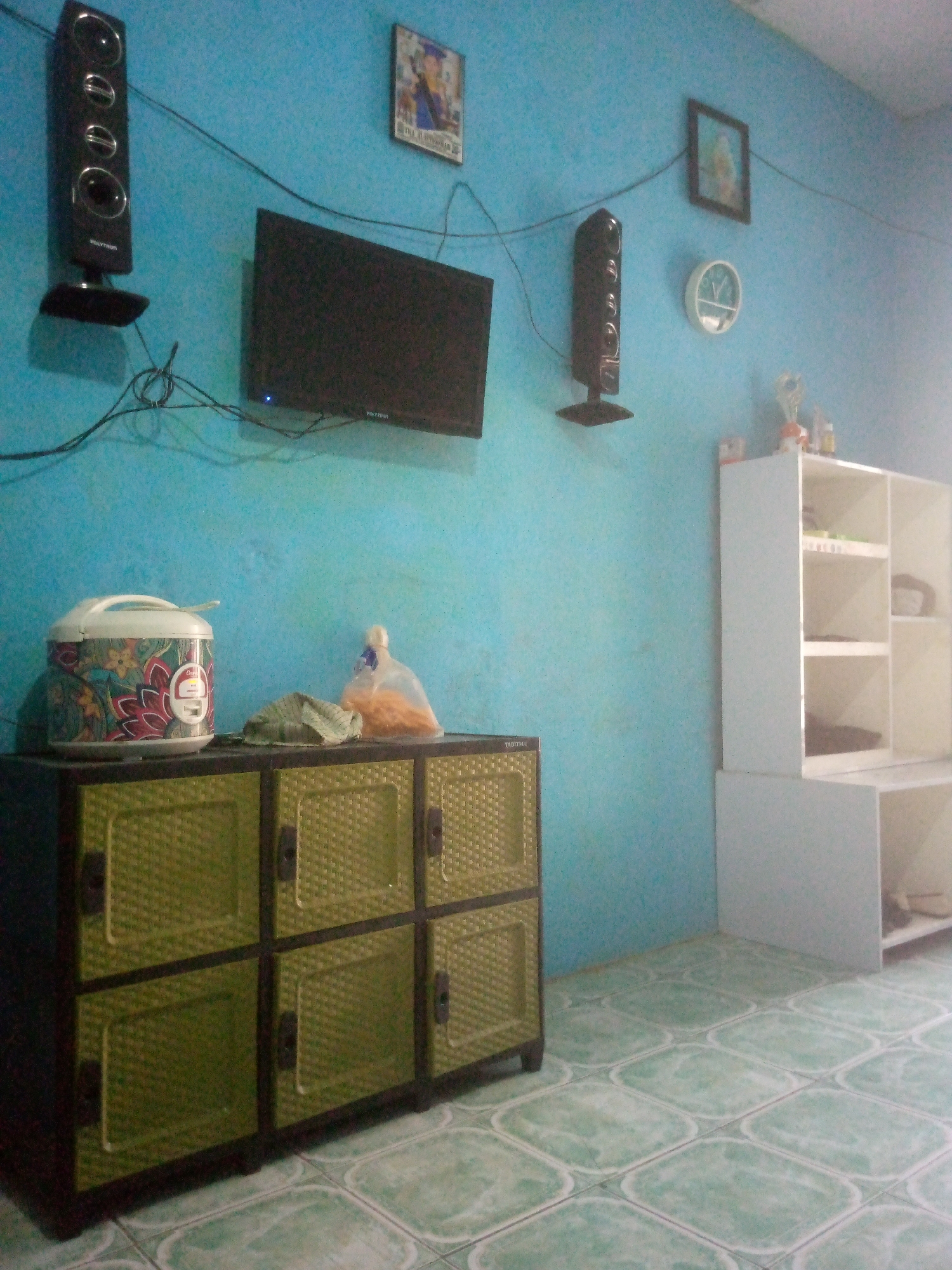 The image depicts an indoor scene, likely a living room or a multipurpose room, with a vibrant turquoise wall serving as the backdrop. To the left, a tall, black speaker is mounted on the wall, positioned above a flat-screen television. The television is also mounted on the wall, with wires extending downwards. To the right of the television, a framed picture hangs on the wall, depicting a person in academic attire. Below the picture, a white circular clock is visible. Further to the right, a white shelving unit stands against the wall, containing several compartments.
In the foreground, a green woven cabinet with multiple drawers and doors is situated. On top of the cabinet, a white rice cooker with a colorful floral pattern is placed, along with a transparent plastic bag filled with food items and a green cloth. The floor is tiled with a pattern of light green and white squares. The overall impression is of a functional, lived-in space. The image does not contain any people. The presence of a rice cooker suggests it may be a residential area. The lighting appears to be natural, possibly from a window not visible in the frame, contributing to a slightly dim atmosphere.