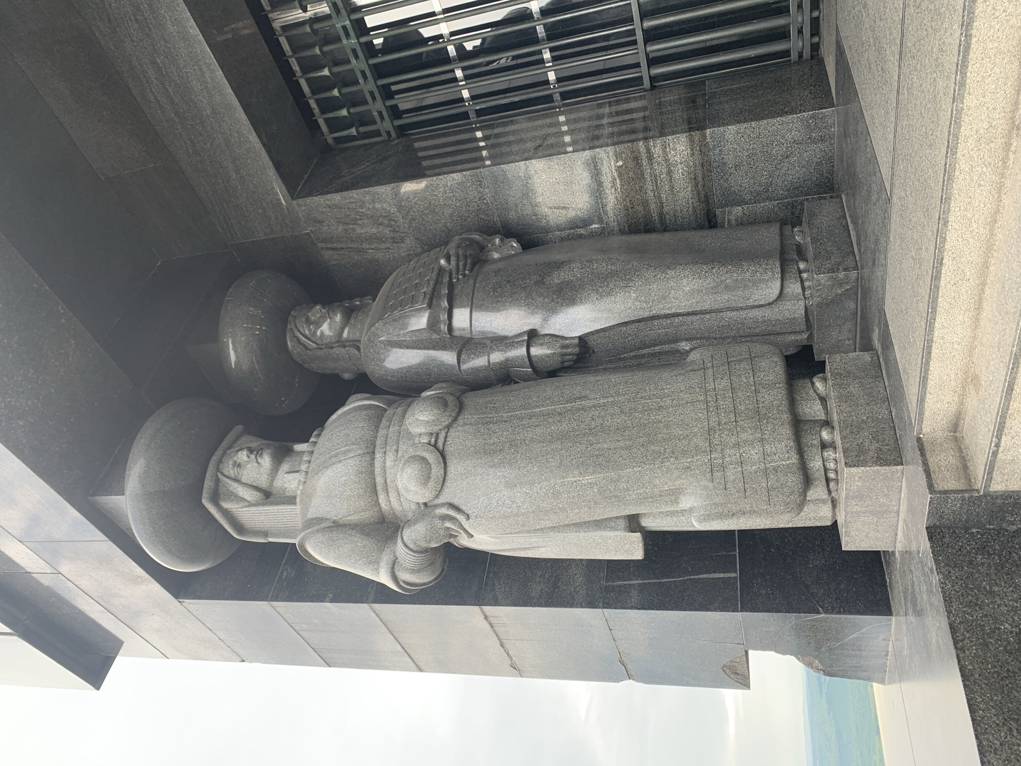 This image shows two gray stone statues of figures in a recumbent position. They appear to be lying on a raised stone platform or structure, possibly part of a monument or memorial. The figures are dressed in what appears to be robes or ceremonial clothing. They have rounded headwear. The statues are indoors, placed within what seems to be a memorial or mausoleum. In the background, there are dark, possibly metal, grilles or bars. The foreground reveals the outside and a hint of trees or nature.
Based on visual similarities to existing images and contextual clues, the location is likely the Memorial Park of Jajinci, which is located in Vozdovac Urban Municipality, Serbia.