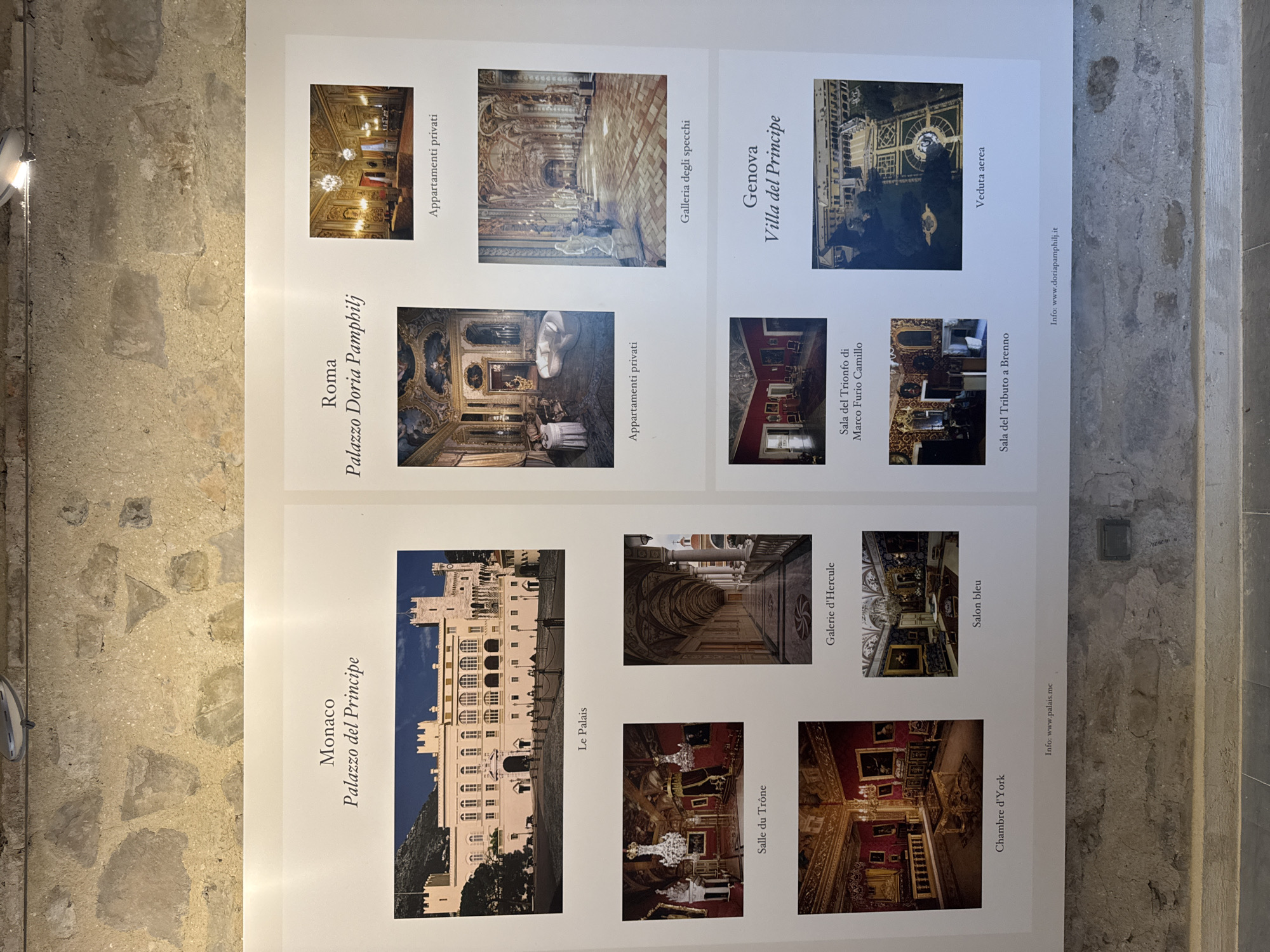 The image is a photograph of a museum exhibit displayed on a wall with a rough, stone texture. The exhibit consists of several framed images and text, organized into three distinct sections labeled "Monaco," "Roma," and "Genova." The "Monaco" section features a large photograph of a palace exterior with the caption "Le Palais," followed by smaller images of interior rooms labeled "Salle du Trône," "Galerie d'Hercule," "Chambre d'York," and "Salon bleu." The "Roma" section displays a photograph of an opulent interior room with the caption "Appartamenti privati," and another image of a grand hallway labeled "Galleria degli specchi." Below these, there are images labeled "Sala del Trionfo di Marco Furio Camillo" and "Sala del Tributo a Brenno." The "Genova" section shows a photograph of a building with manicured gardens from an aerial perspective, captioned "Veduta aerea," and the text "Villa del Principe." Website information is provided at the bottom of each section. There are no people visible in the exhibit or its surrounding environment. The exhibit is mounted on a white surface against a backdrop of rough-hewn stone walls, suggesting an indoor display space. The lighting appears to be artificial, casting a soft illumination on the exhibit. The city of Dolceacqua, Italy, is not depicted or mentioned in the image or its accompanying text.