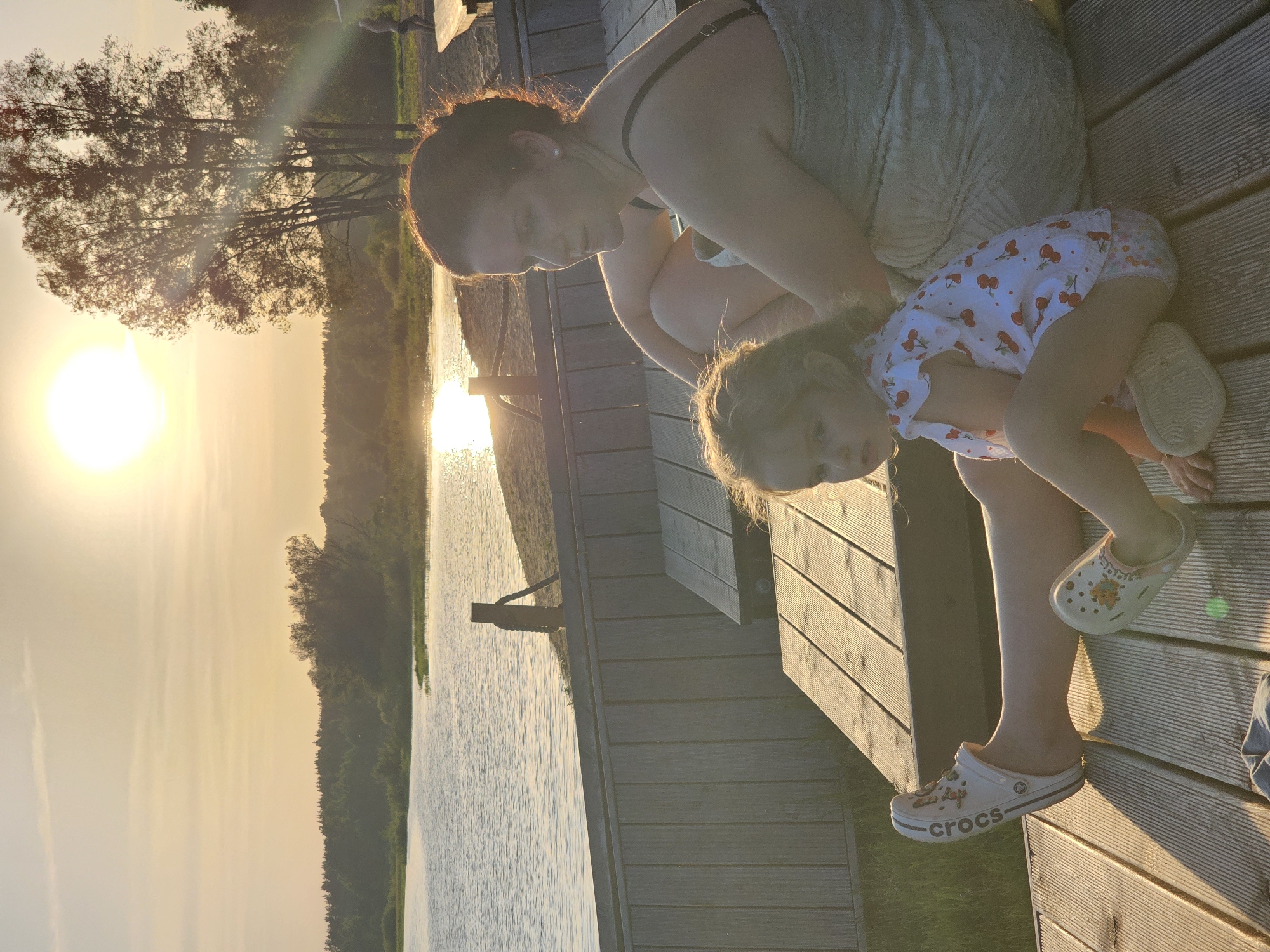 On a wooden deck near a body of water reflecting the setting sun, a woman sits with a young child. The child, wearing a white outfit with red cherry patterns and white Crocs shoes, leans against the woman's leg. The woman, wearing a light-colored dress, is looking down at the child. The backdrop features trees and the sun casting a golden glow over the water and sky. The image may be taken in Мышкино, Russia, given the prompt's reference.