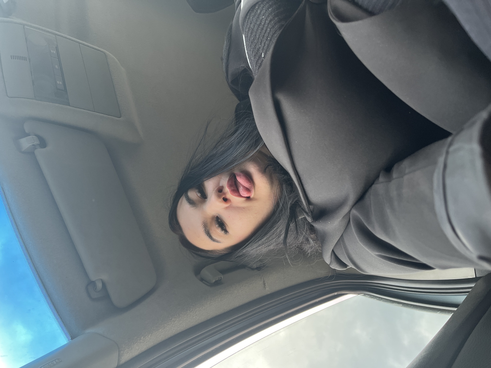 A low-angle shot captures a person inside a vehicle, likely a selfie taken looking upwards. The subject, a young person with long, dark hair, light skin, and dark eyebrows, is making a playful face by sticking out their tongue. They are wearing dark clothing, possibly a jacket over a ribbed top. Their head is oriented upside down in the frame relative to the car's interior.
The vehicle's light-colored headliner and a detailed roof console are prominent in the background. The console features various rectangular panels and buttons, with some faint, unreadable text or symbols visible. A sun visor is partially visible on the left side of the frame, and another is folded up near the center. Through the vehicle's windshield and what appears to be a sunroof or side window, a bright blue sky with scattered light clouds is visible, indicating it is daytime. The scene suggests a lighthearted moment within a car in Chaikovsky, Russia.