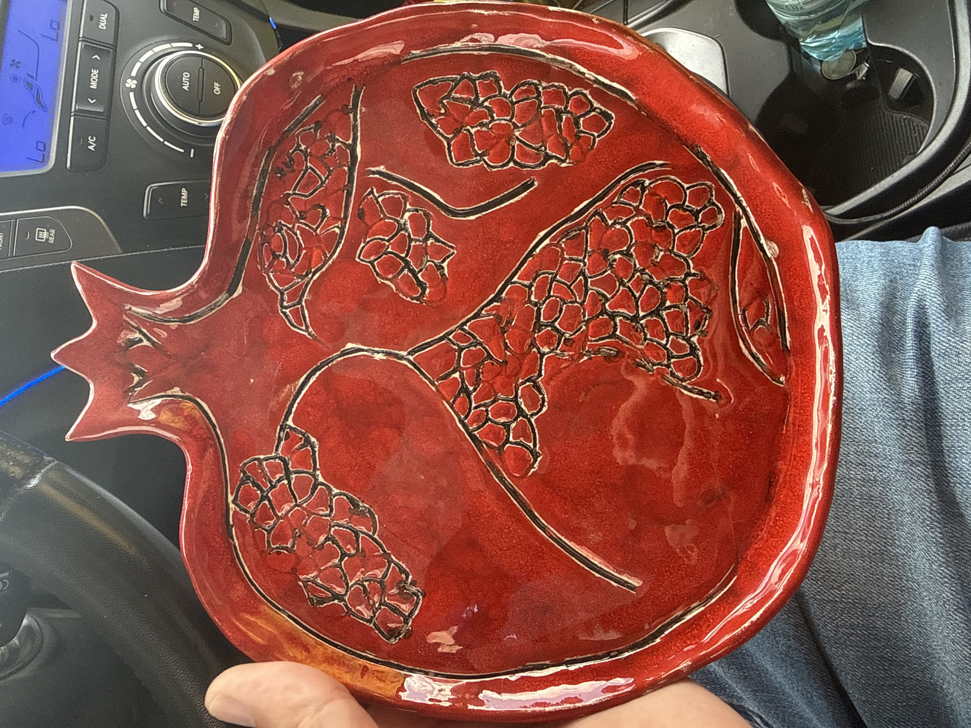 A red, pomegranate-shaped ceramic dish is held in a person's hand. The dish has raised detailing representing pomegranate seeds, outlined in black. The background shows the interior of a car, including a steering wheel and a car's dashboard with various buttons and a digital display. This scene does not provide any information that would allow for determining its location in the city of Novy Urengoy, Russia.