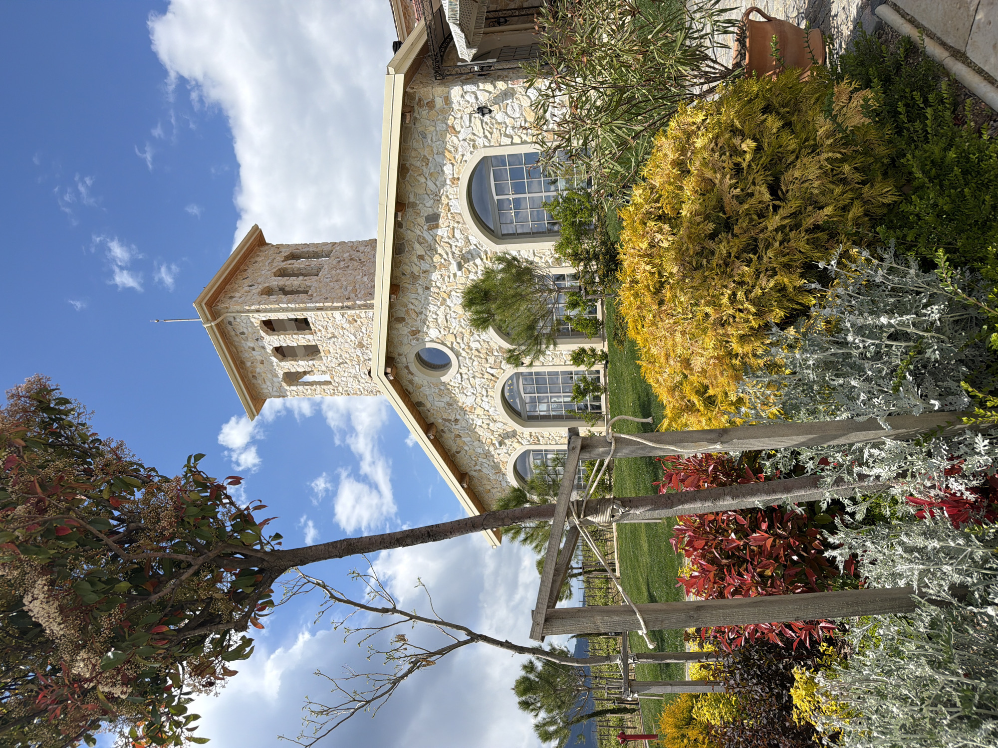 The image shows a stone building with arched windows and a tower under a blue sky with white clouds. The building is surrounded by green grass, bushes, and colorful flowerbeds. In the foreground, a wooden fence is visible. The photo appears to be taken in Kocherinovo, Bulgaria.