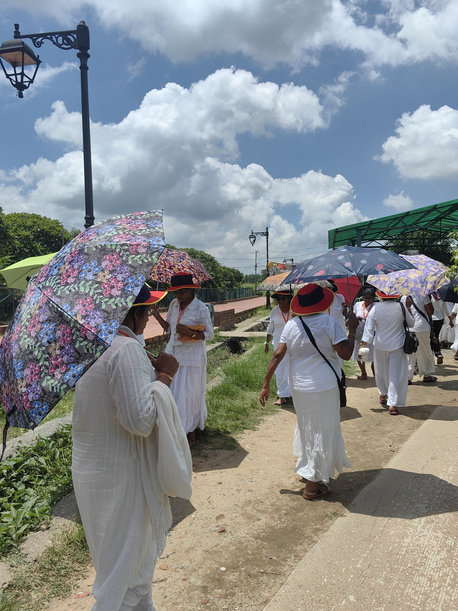 In Lumbini Bazaar, Nepal, a group of women dressed in white attire walk along a dirt path. Many are holding colorful umbrellas to shield themselves from the sun. The sky is bright blue with fluffy white clouds. A lamppost stands tall on the left side of the image, and a green structure is visible in the background. The women appear to be walking together, some engaging in conversation as they move.