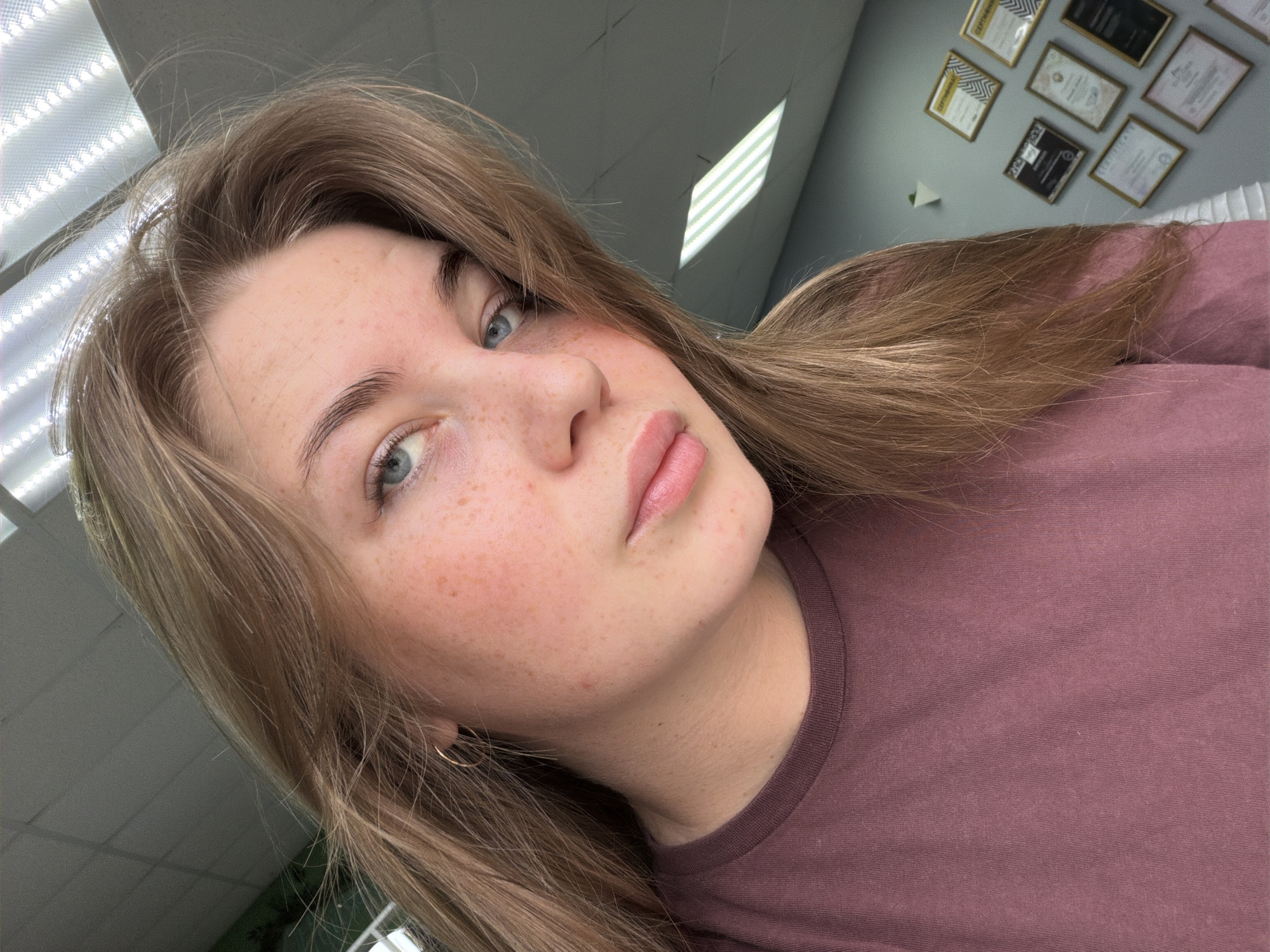 A close-up, high-angle selfie shows a young Caucasian woman with long, light brown hair and blue eyes. She has fair skin with a smattering of freckles across her nose and cheeks. She is wearing a mauve t-shirt. The background suggests an indoor environment, possibly an office or classroom, with fluorescent lighting visible on the ceiling and framed certificates displayed on a gray wall. The image is taken in Chaikovsky, Russia.