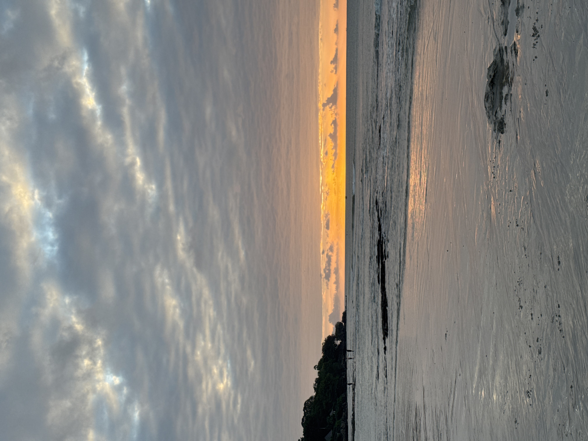 A wide-angle, eye-level shot captures a serene beach scene in Kuta Selatan, Indonesia, during a sunrise. The sky is a canvas of soft, muted colors, with bands of gray and pale orange clouds stretching across the frame. The sun, a bright orange orb, peeks through the horizon, casting a warm glow on the water and sky. Gentle waves roll onto a wet sand beach, leaving behind intricate patterns from the receding tide. In the distance, a lush green, tree-covered cliff juts out from the coastline. A few silhouetted figures, appearing small against the vastness of the beach, are visible near the water's edge. The overall atmosphere is tranquil and peaceful, evoking the calm beauty of a tropical morning.