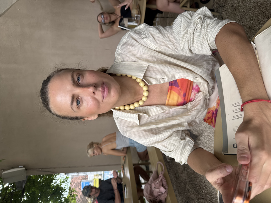 The image captures an outdoor or semi-outdoor cafe scene in Uluwatu, Indonesia. A fair-skinned woman with light blue eyes and light brown hair, pulled back, is positioned in the foreground, looking directly at the camera with a subtle smile. She wears a cream-colored, vertically striped, long-sleeved button-down shirt unbuttoned over a vibrant orange and pink patterned strapless top. A chunky, light yellow beaded necklace adorns her neck. Her left hand holds a rose gold-colored smartphone, while her right hand rests on a light-colored tabletop next to an open menu. A thin red string bracelet is visible on her right wrist.
In the mid-ground to the left, a man wearing a dark t-shirt and a camouflage baseball cap is seated at a wooden picnic-style table, partially obscured, appearing to look down at the table. Next to him, a blonde-haired woman in a light gray tank top and denim shorts is seated, facing left and away from the camera. Food items are visible on their table. An outdoor speaker is mounted on the light-colored wall above them, with green foliage and a street scene visible further in the background.
To the right, another blonde-haired woman, wearing sunglasses and a black strapless top, is seated at a separate table, leaning back with her hands positioned behind her head. A drink with a straw is on her table. The background wall is a plain light tan color. The ground beneath the tables appears to be covered in gravel or a similar material.