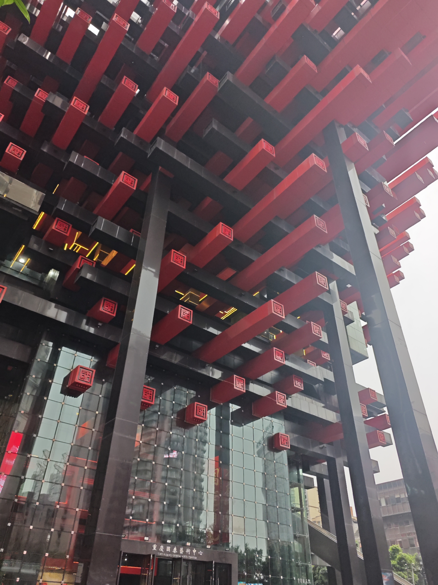 The image shows a low-angle view of the Raffles City Chongqing complex, located in the Yuzhong District of Chongqing, China. The structure features a modern design with a combination of glass and steel. The building's exterior is characterized by a grid-like pattern of red and black rectangular elements extending upwards. Some of these elements are adorned with small red squares, each featuring a Chinese character or symbol. The glass facade of the building reflects the surrounding urban environment. The sky appears overcast. The bottom of the image displays the entrance area with signage in Chinese and English reading "CHONGQING GUOTAI ARTS CENTER." A few people are visible near the entrance.