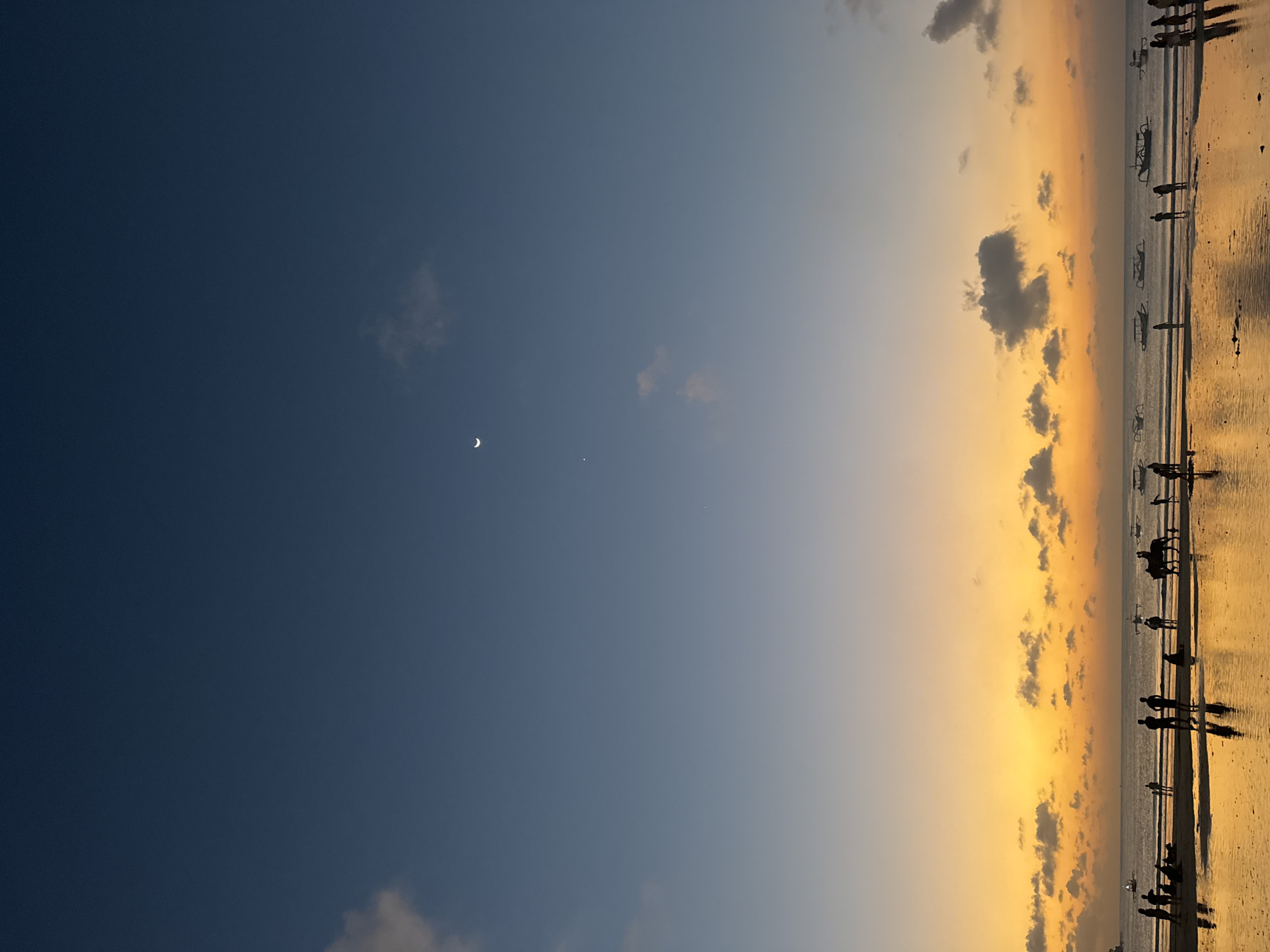 This image depicts a sunset scene at Kuta Selatan, Indonesia, featuring a beach with numerous people and several boats in the ocean. The sky transitions from a deep blue at the top to a vibrant orange and yellow near the horizon, with scattered clouds. A crescent moon is visible in the upper portion of the sky.
In the foreground, the wet sand reflects the colorful sky, with several silhouetted figures scattered across the beach. Some individuals are standing, while others are seated. To the right, a person is riding a horse.
The ocean is calm, with multiple traditional Balinese boats, locally known as jukung, anchored or sailing on the water. The silhouettes of the people and boats are stark against the bright sky and its reflection. The overall atmosphere is serene, capturing the beauty of a tropical sunset.