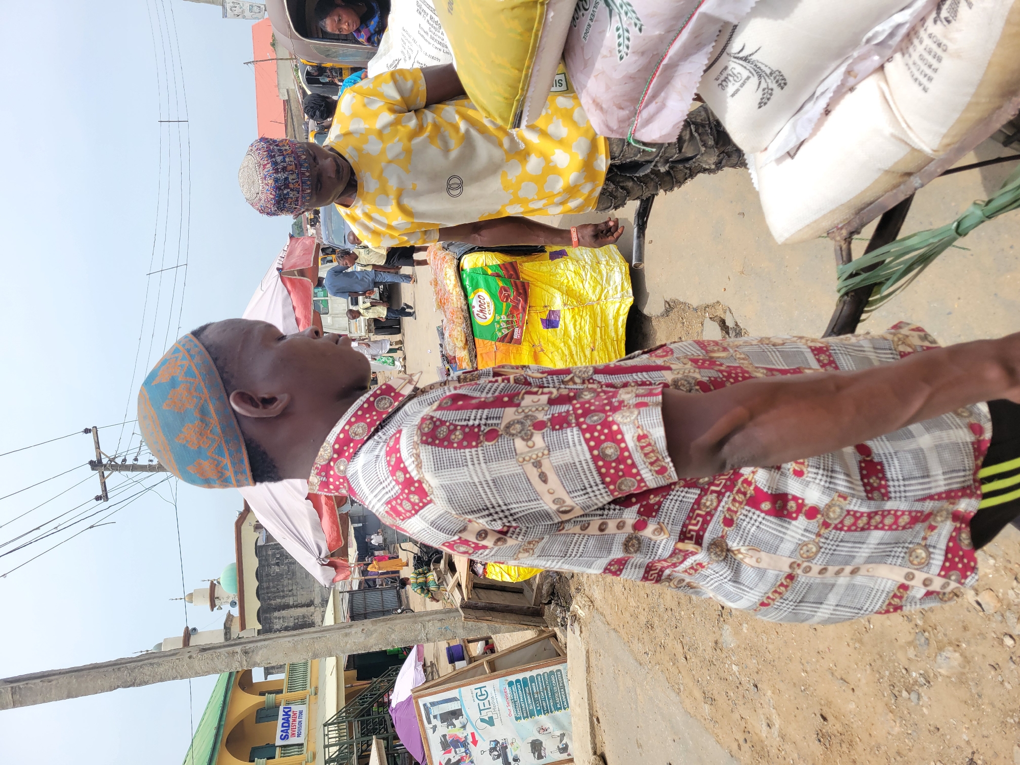 Here's a detailed description of the image:
**Setting:** The scene appears to be in a bustling commercial area, possibly a market, in Suleja, Nigeria. Elements indicating this include the presence of street vendors, commercial signage, and a mix of building styles. The ground surface is a combination of pavement and dirt.
**People:** Two men are prominently featured in the foreground. One is wearing a red and white patterned shirt and a blue and orange cap. The other man is wearing a yellow and white patterned shirt and a colorful cap, and he is standing beside sacks of goods that he appears to be selling. Other individuals are visible in the background, suggesting a crowded environment. A woman is riding in a "Keke Napep" (auto rickshaw).
**Objects:**
* Sacks of rice and other provisions, suggesting the selling of food or commodities.
* A cart used to transport the goods.
* Commercial signage for "TECH Medias" (advertising various print and media services) and "SADAKI INVESTMENT PROVISION STORE" indicating types of businesses in the area.
* A packet of "Choco Cure" chocolate product.
* Electrical power lines and poles.
* Buildings of varying construction and condition in the background.
* A mosque with a green dome in the background.
**Actions/Interactions:**
* The people are in a commercial setting and likely engaged in buying or selling. The men in the foreground are likely vendors.
**Environment:**
* The weather appears to be sunny, based on the lighting.
* The scene is likely a busy street corner with pedestrian and vehicular traffic.