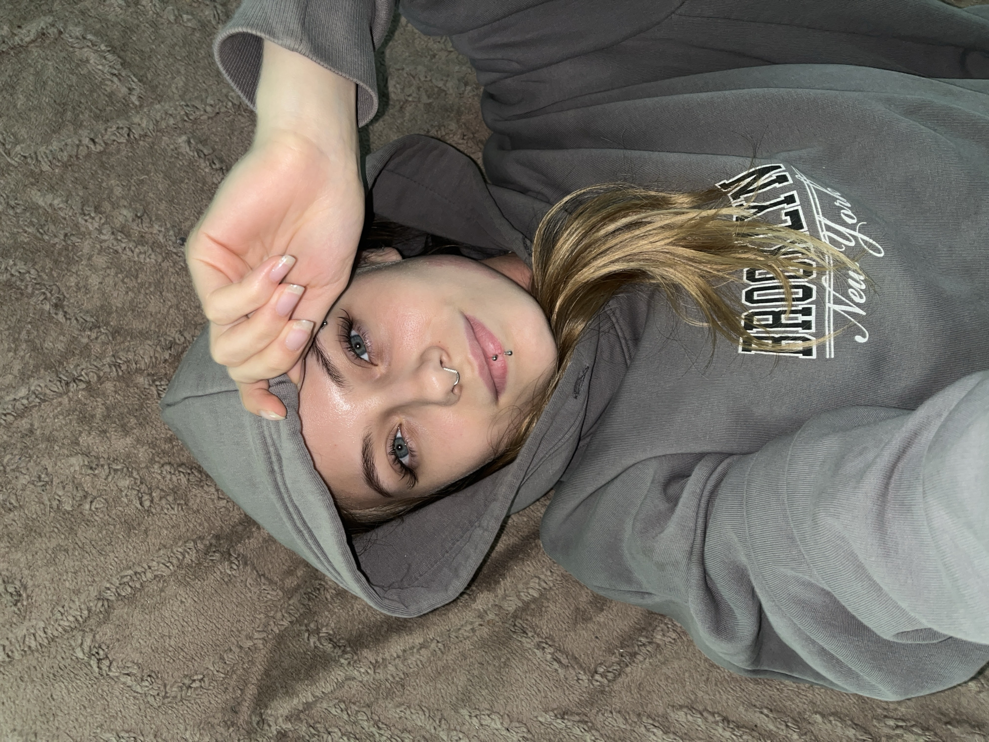 A young woman is lying on a brown, textured surface, possibly a rug or carpet. She is wearing a gray hooded sweatshirt with "Brooklyn New York" printed on the chest. She has light skin, blonde hair, and blue eyes. She has piercings in her nose and lip. Her left hand is raised towards her forehead, partially obscuring her face. The scene appears to be indoors and is lit with a soft, ambient light. The location of Фоки, Russia, is not apparent from the image.