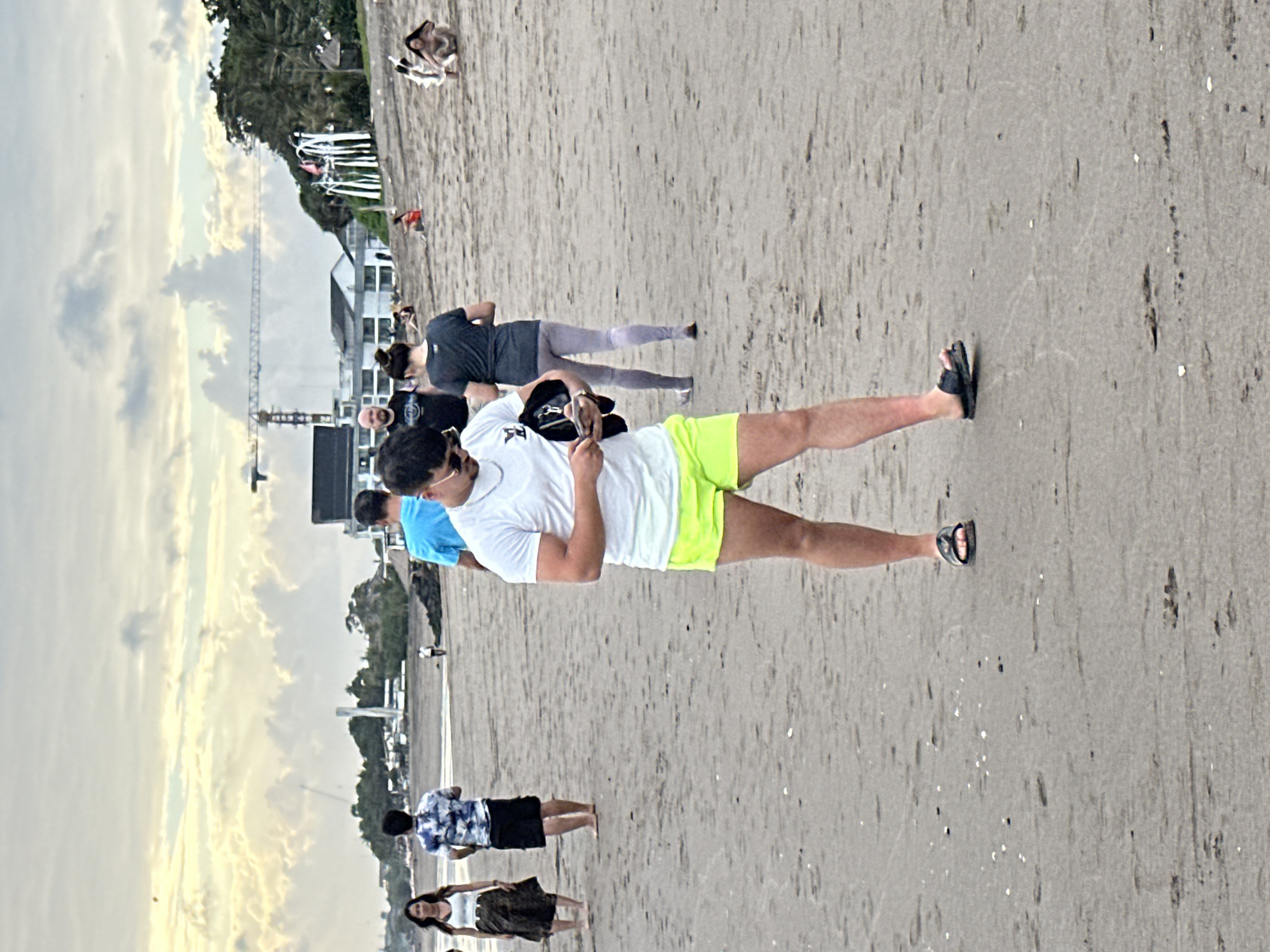 In North Kuta, Indonesia, a group of individuals are present on a sandy beach under an overcast sky. The primary subject, a young man in the foreground, is wearing a white t-shirt, bright yellow shorts, and black flip-flops. He is looking down at his phone, holding a dark-colored jacket or bag in his left hand. Behind him, several other people are walking on the beach. To his left, a woman in a white tank top and a patterned skirt is walking, followed by a man in a blue and white patterned shirt and black shorts. Further in the background, other beachgoers are visible, some sitting and others walking. Buildings and a construction crane can be seen in the distance, suggesting ongoing development in the area. The beach appears to be a public space with light foot traffic.