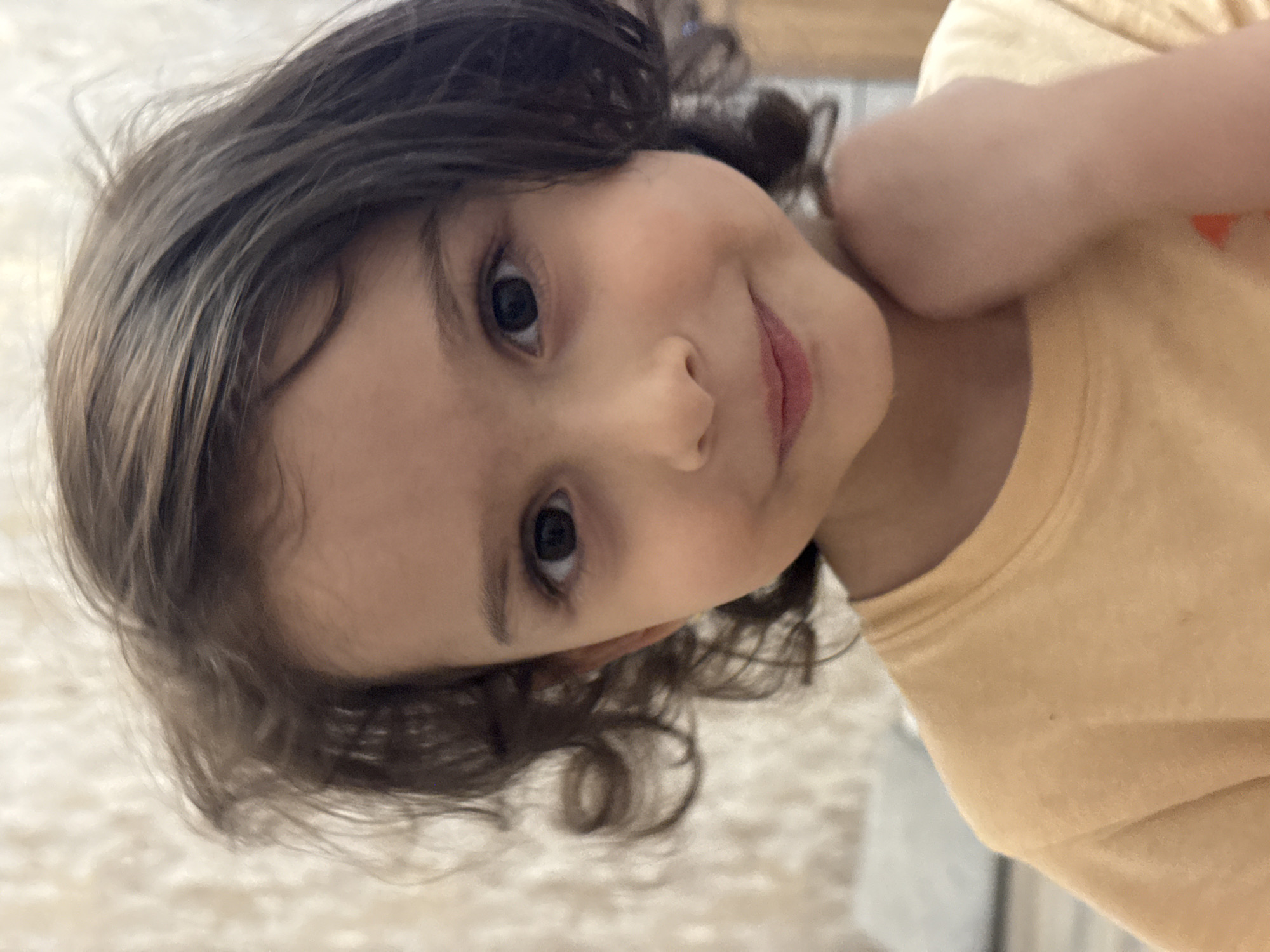 A close-up, eye-level, indoor studio portrait shows a young child, appearing to be of Eastern European or Middle Eastern descent, with curly dark brown hair and fair skin. The child is wearing a plain, light orange t-shirt. The child's head is slightly tilted to their left, and their gaze is directed towards the camera. Their right hand is raised and touching their neck, near their jawline. The background is a textured, light-colored wall with subtle vertical lines. The lighting is soft and appears to be coming from the front. The image is sharp on the child's face, with a shallow depth of field blurring the background. The image appears to have been taken in Baku, Azerbaijan, given the style of the textured wall and the overall aesthetic.
